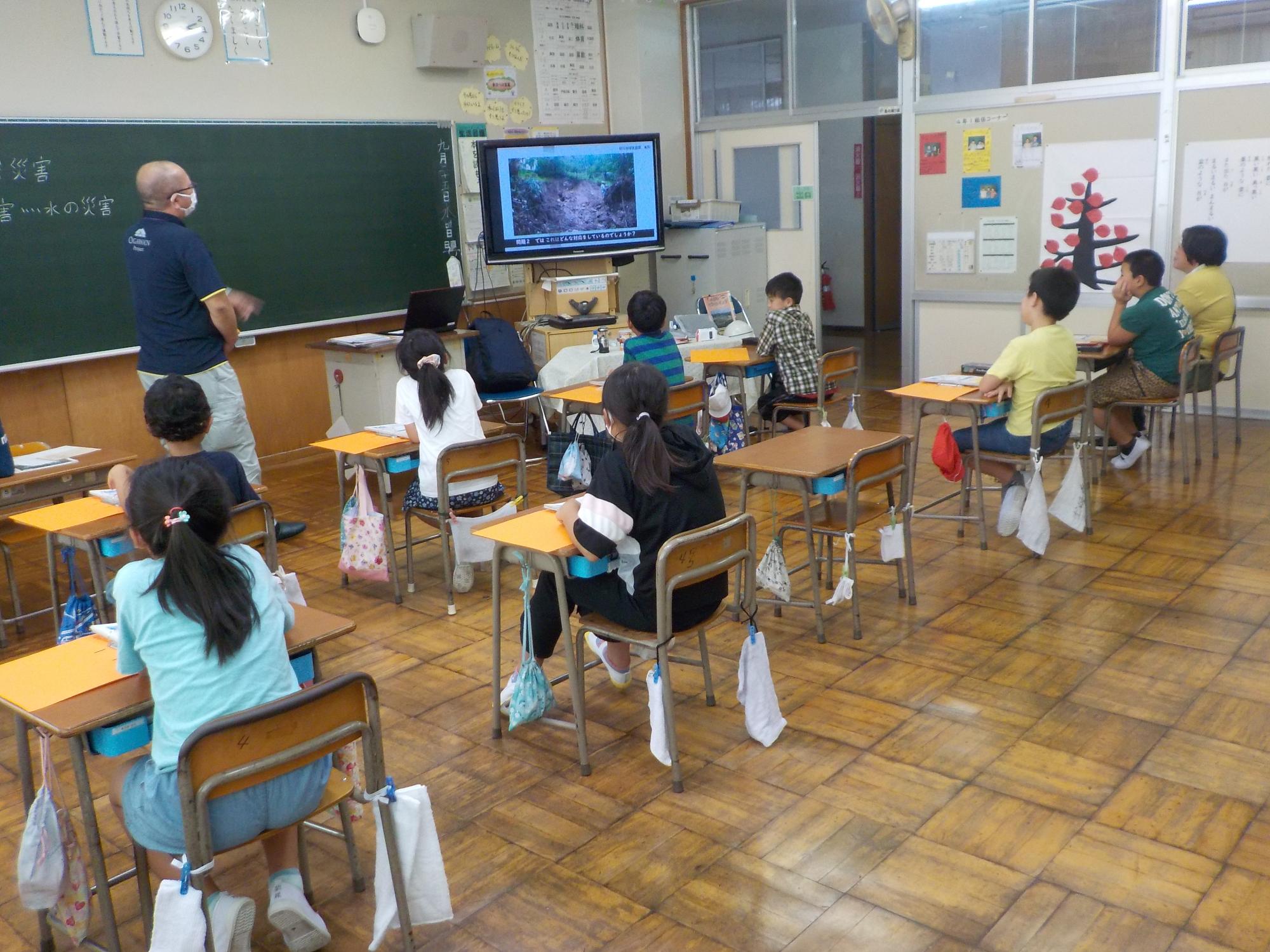 黒板の横に設置されたモニターを机に座りながら子供たちが見ている前に講師の男性が立っている写真