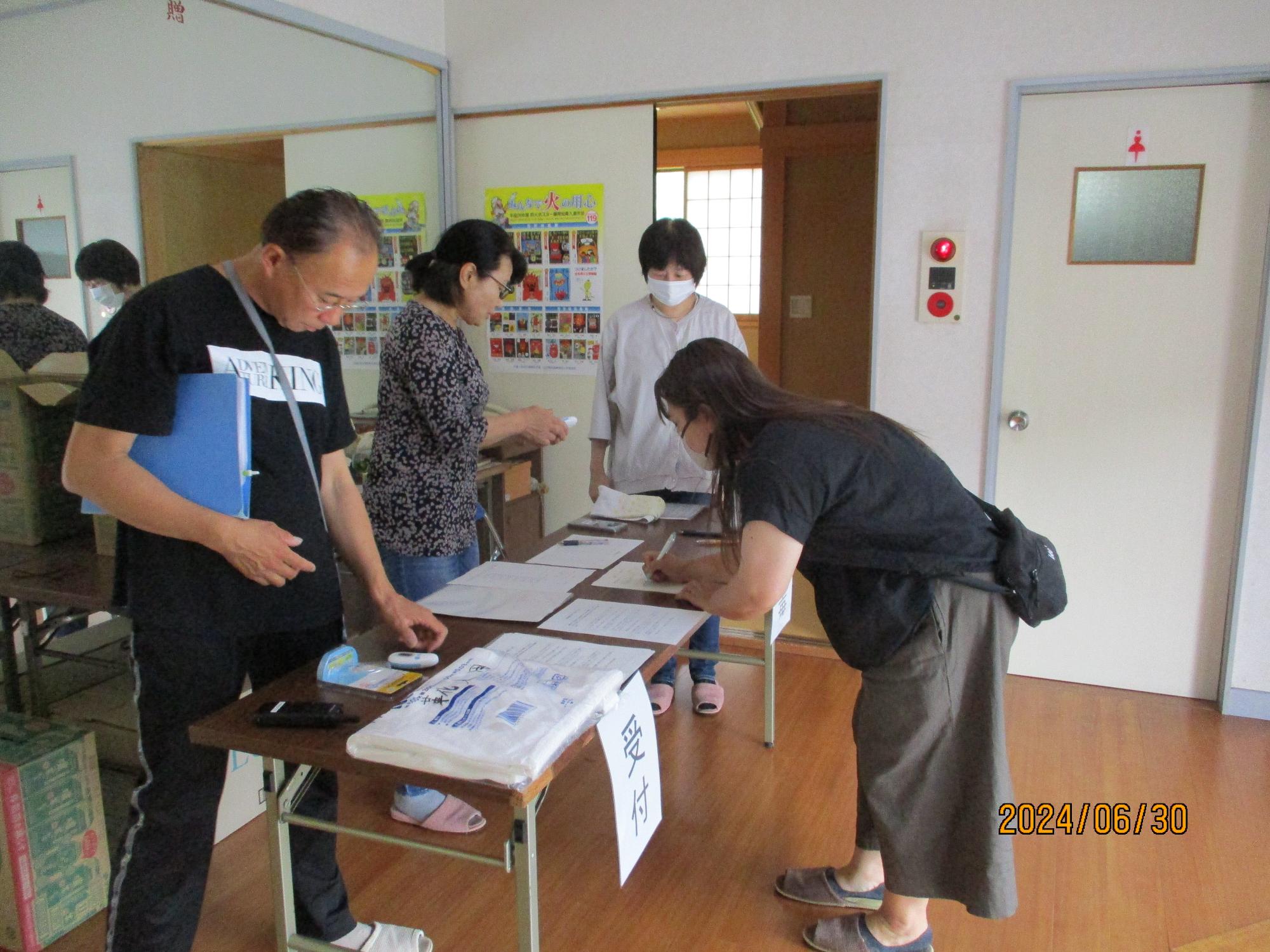 男性と女性2人が立っているテーブルを挟んで1人の女性が避難者カードに記入する様子を横から撮影した写真