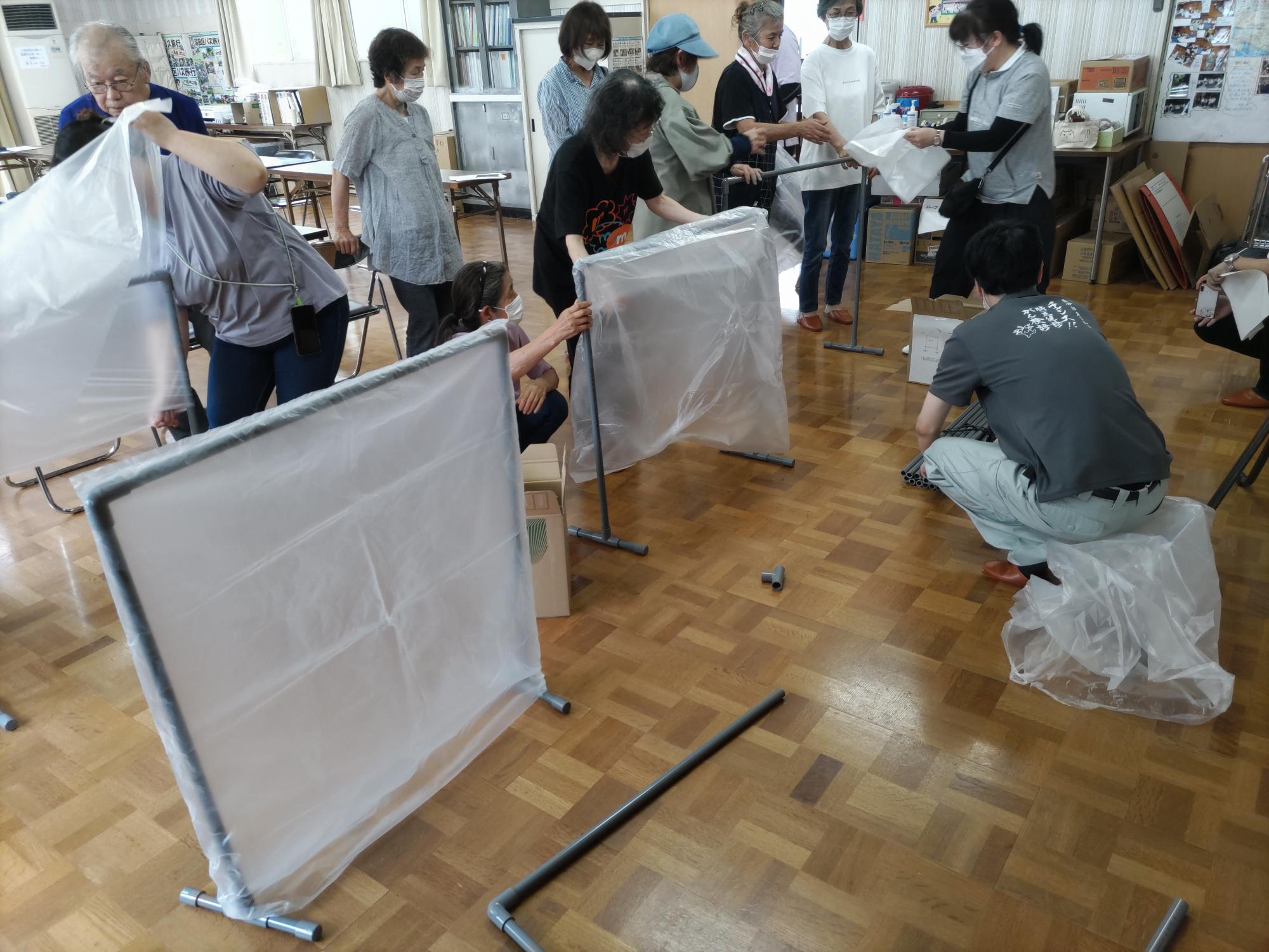 深田区民センターのフローリングの床でパーテーションに袋を掛けたり組み立てている参加者の方々の様子を撮影した写真