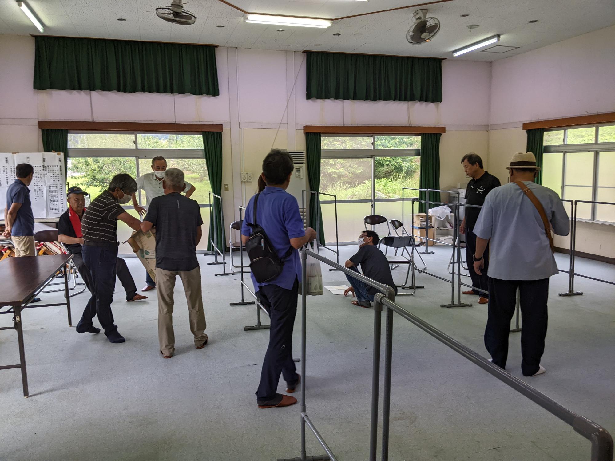 上勝呂会館の広いフロア中央付近で参加者の方々がパーテーションの区画について話している様子を後方から撮影した写真