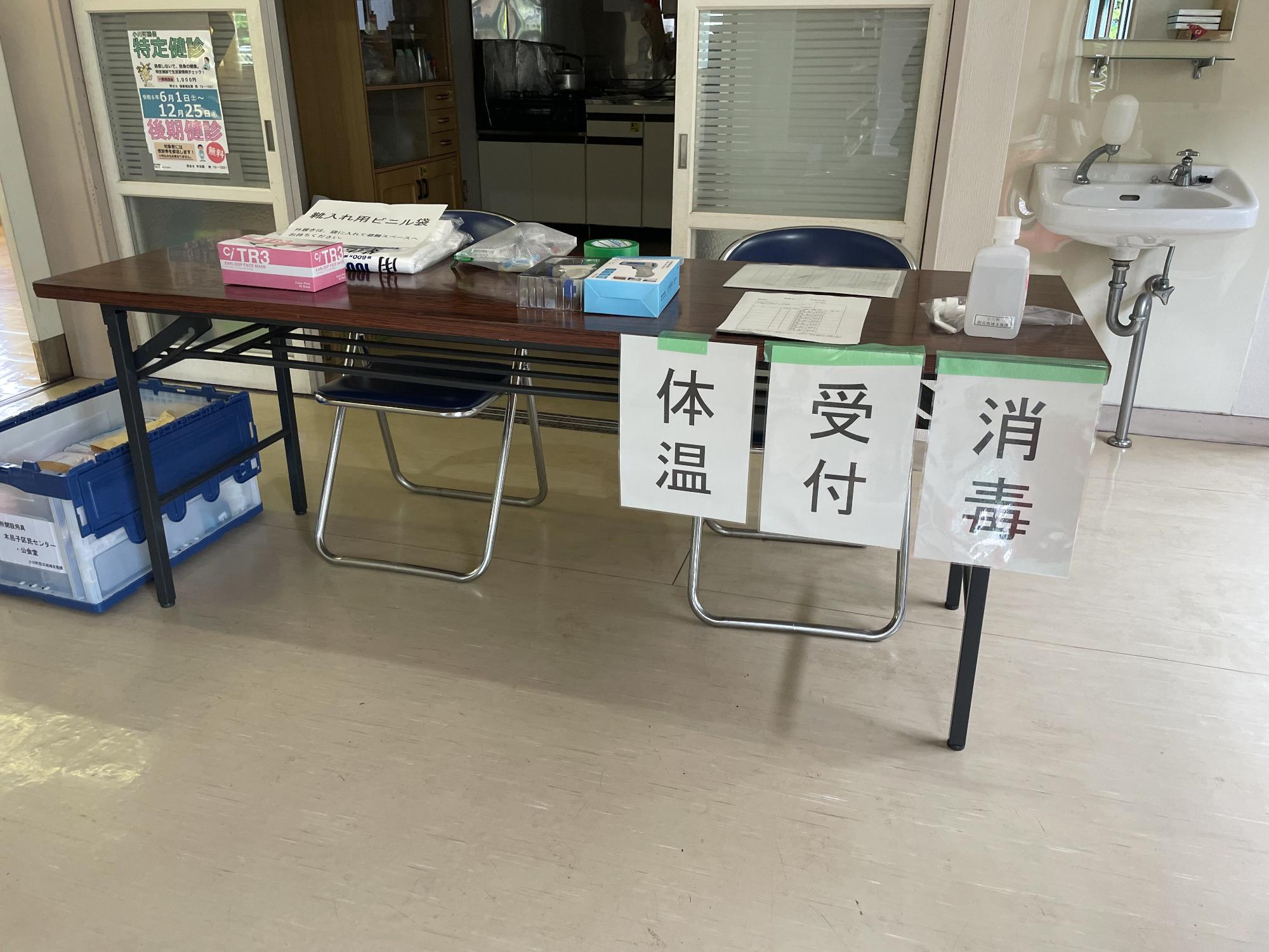 長机に2脚のパイプ椅子が設置され消毒、受付、体温の紙が貼られた避難場所受付の様子の写真