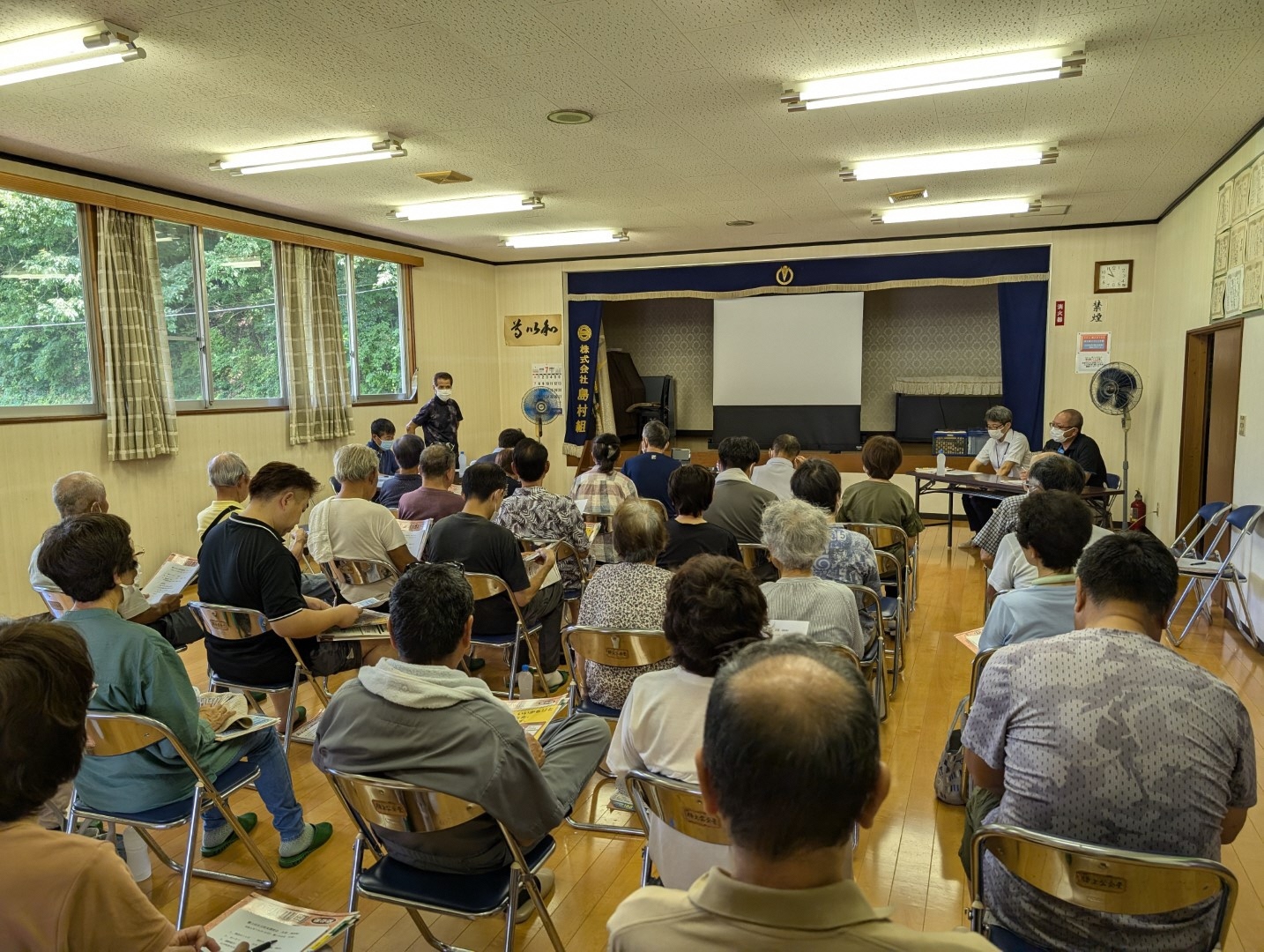 スクリーンが前方にあり、縦5列に並んだパイプ椅子に座る多くの参加者の方々の後姿を撮影した写真