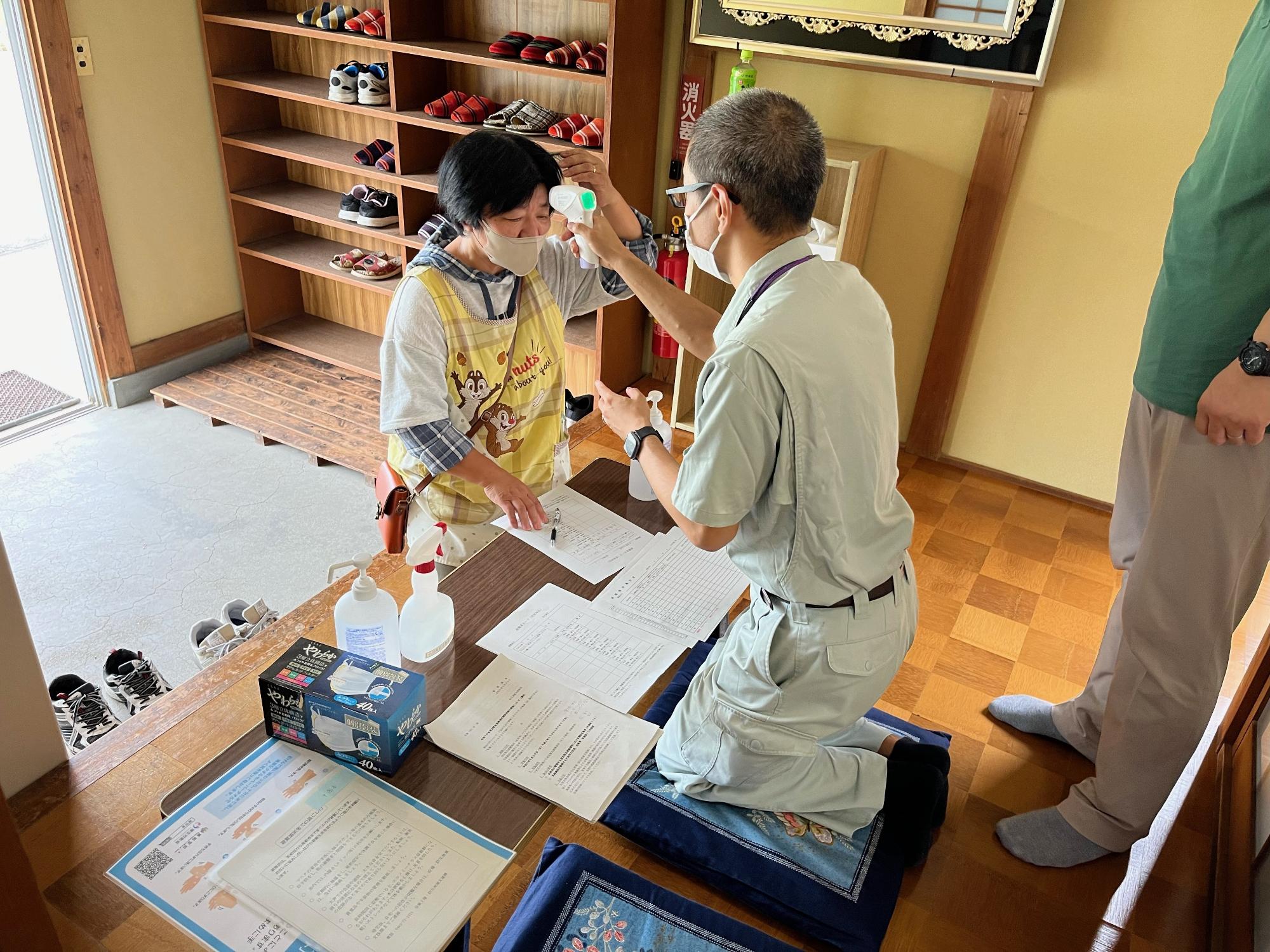 中高谷公会堂の玄関で体温計を持った男性職員がエプロン姿の女性の額に当て体温測定している様子を撮影した写真