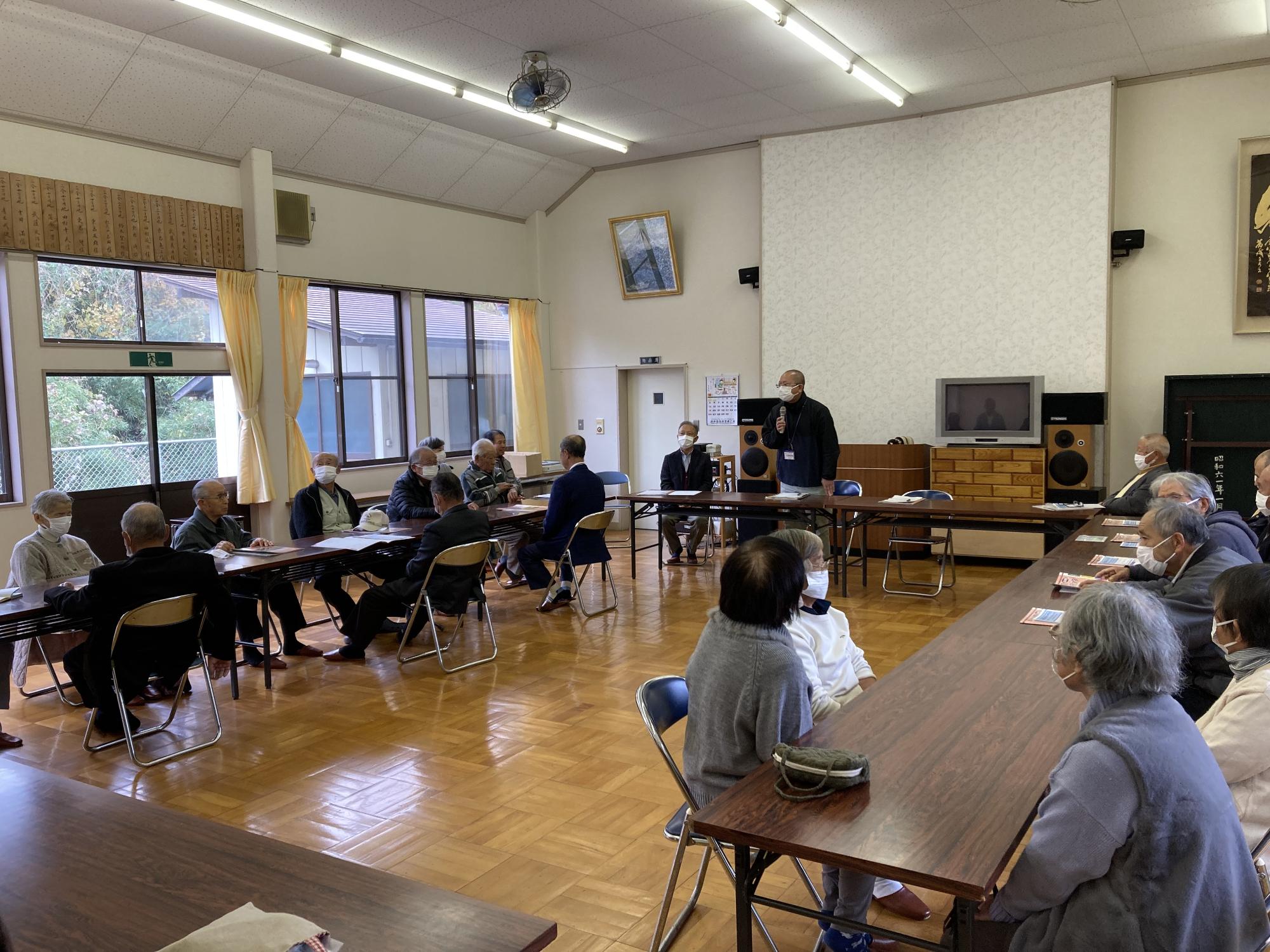 コの字型に設置されたテーブルと椅子に向かい合って座るっている参加者の方が前方に立つ男性の説明を聞いている様子を後方から撮影した写真