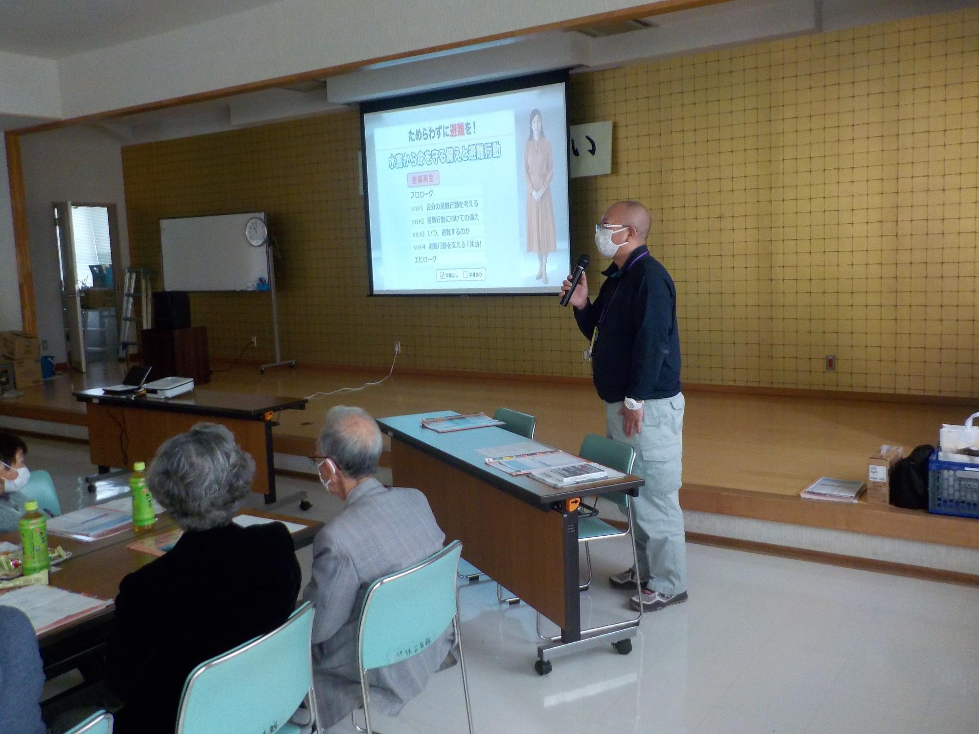 部屋の中でマイクを持った男性が防災講座の参加者たちの前に立ちながら話をしている写真