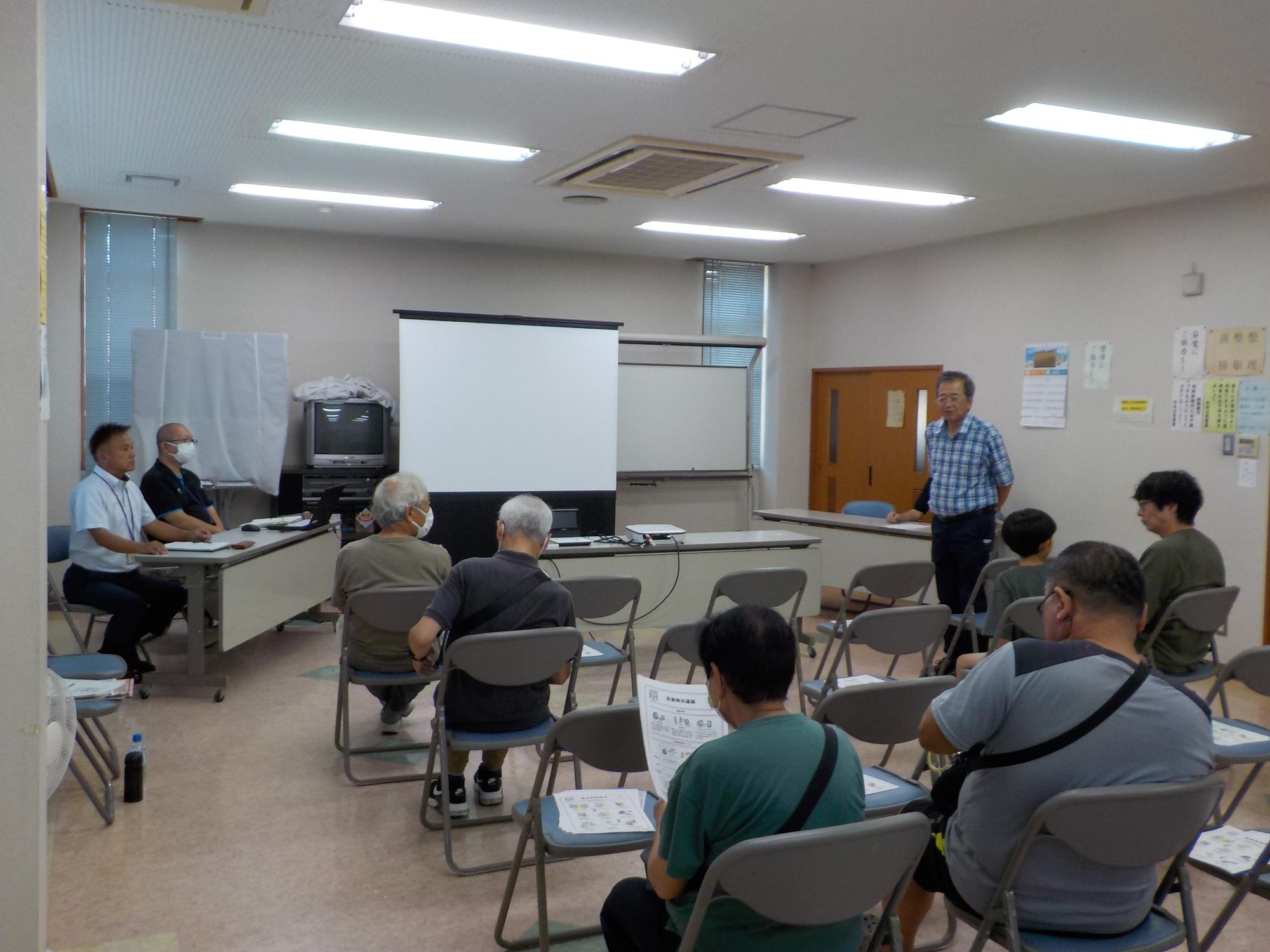 スクリーンとパイプ椅子に座る参加者の方々の間に立ってあいさつをしている区長の様子を撮影した写真