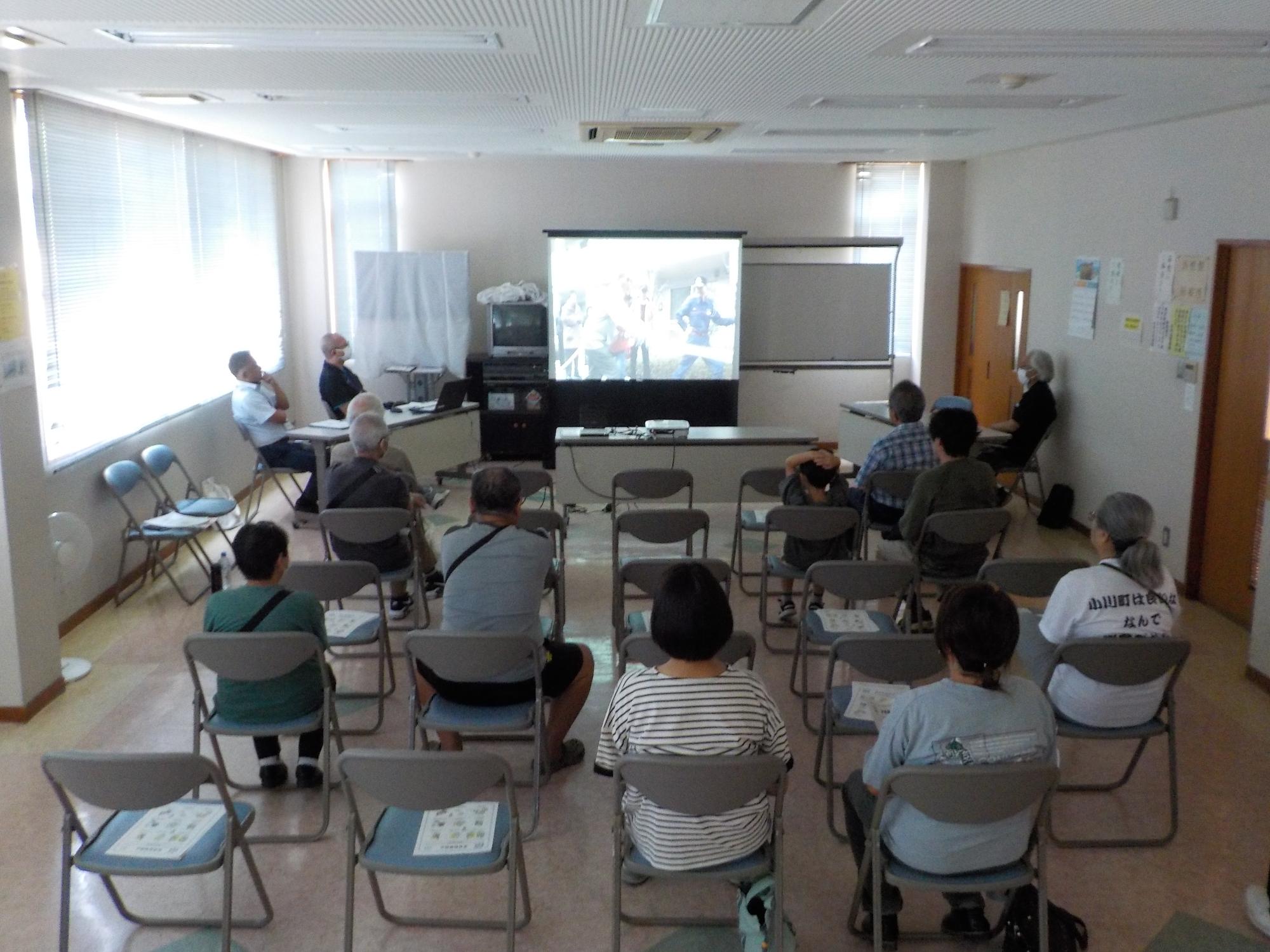 前方のスクリーンに映し出された映像を熱心に見ているパイプ椅子に座った受講者の方々の後姿を撮影した写真