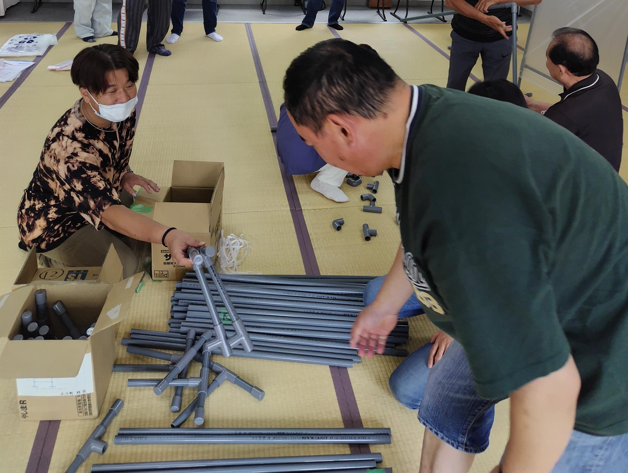 畳の部屋で箱からパーテーションの部品を取り出して手渡そうとする男性と受け取ろうとしている男性の様子を撮影した写真