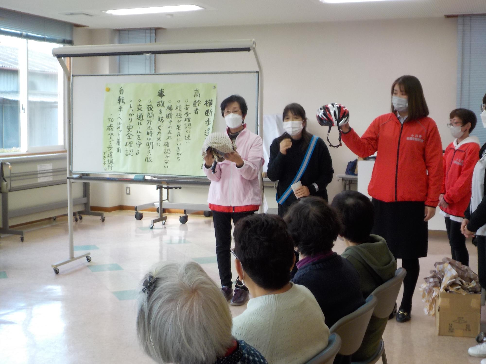 ホワイトボードの横に立ってヘルメットを手にした女性2人と反射材を着けた母の会のメンバーと説明を聞いている参加者の方々の様子を撮影した写真