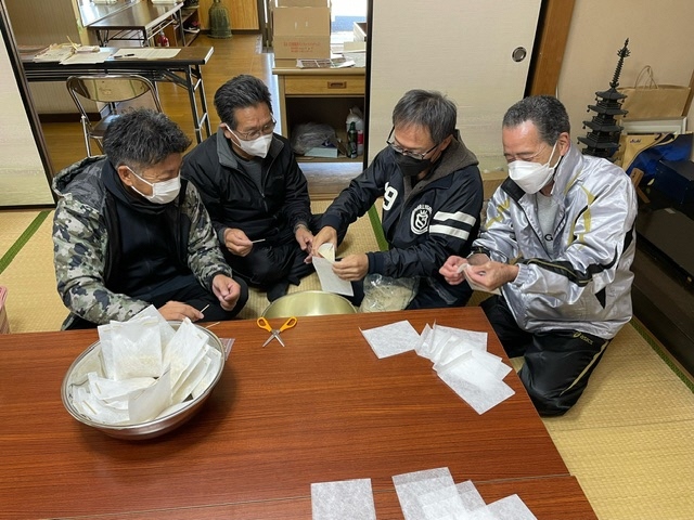 4人の男性が畳の上に座り白い半透明の小袋に物を詰めている写真