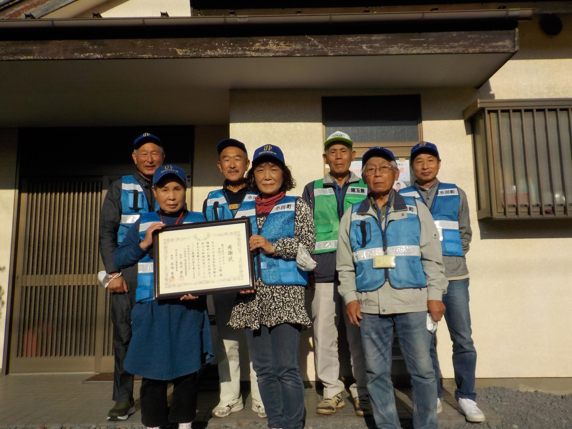 複数の男女が建物の前で感謝状の賞状を手に持ちながら横並びに並んでいる写真