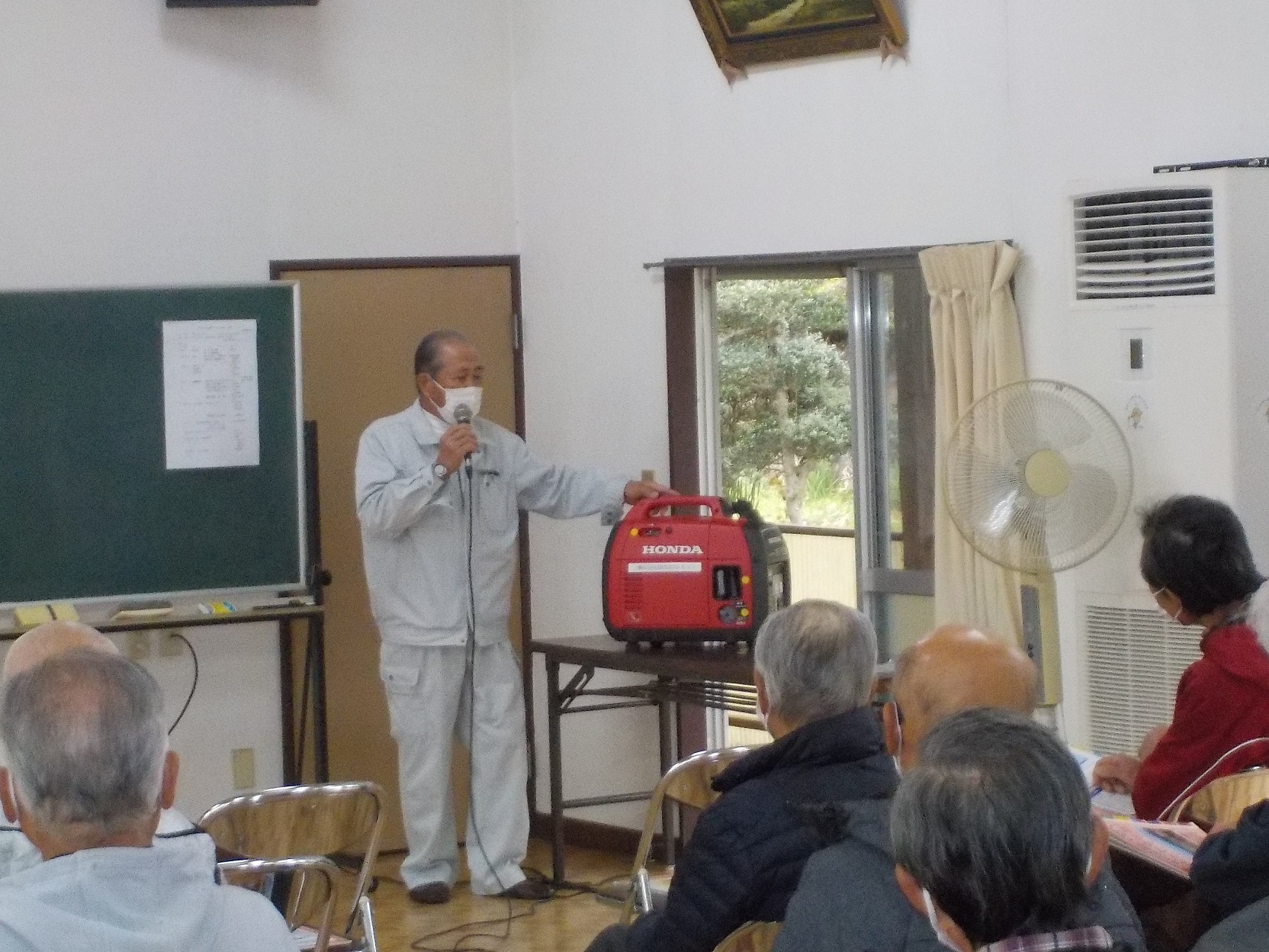 屋内でマイクを持った男性が参加者たちを前に赤い発電機の使い方を説明している写真