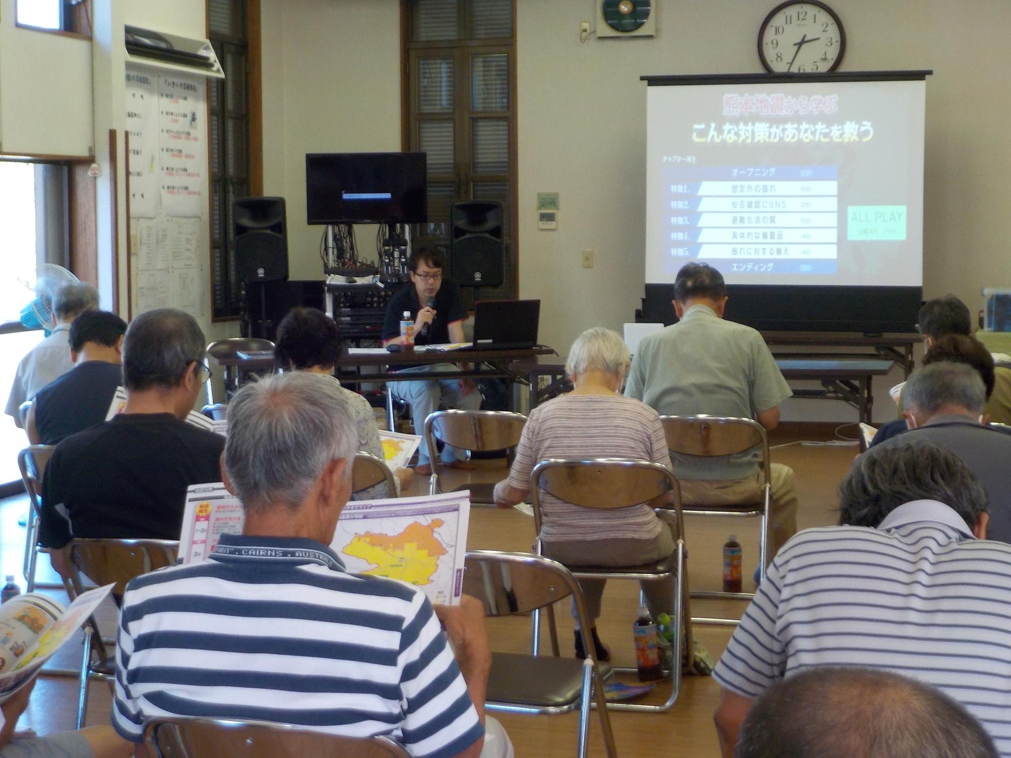 前方のスクリーンに映し出された映像の説明に耳を傾けながら手に持った資料を見ているパイプ椅子に座った参加者の方々を後方から撮影した写真