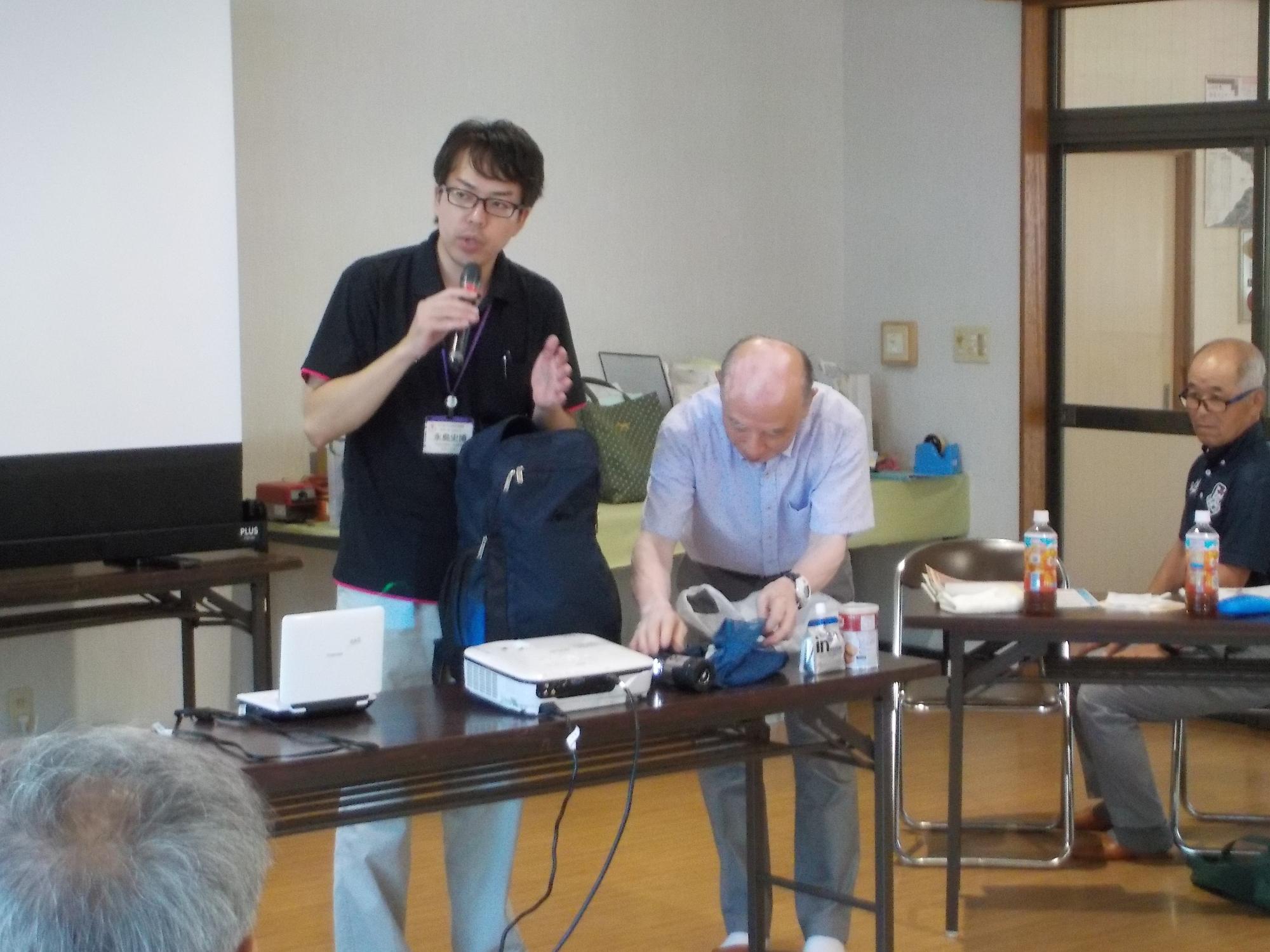 テーブルの前に立つ2人の男性が防災リュックの中身を出して説明している様子を撮影した写真