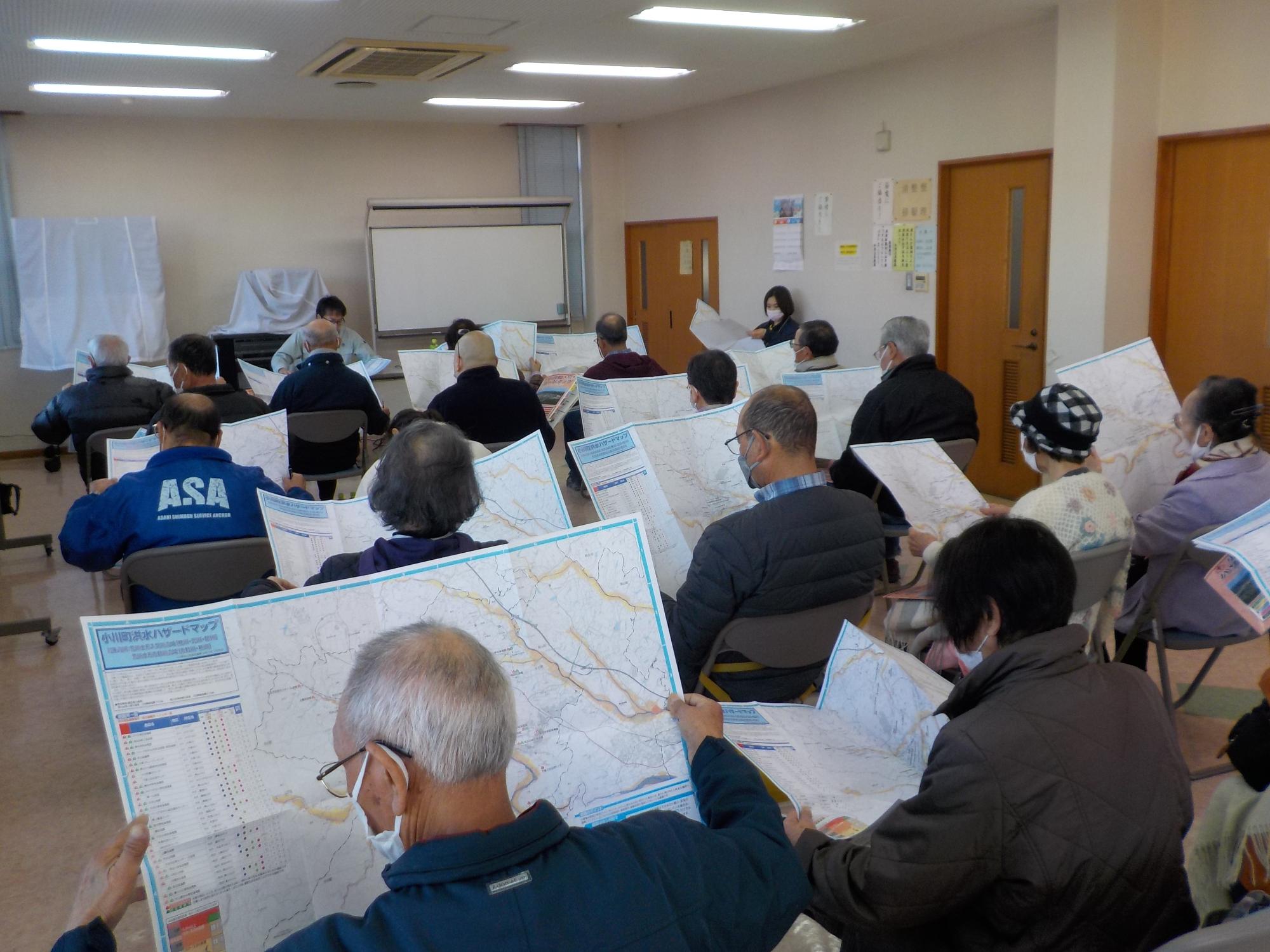 前方のホワイトボードに向かってパイプ椅子に座った参加者の方々が手に持った地図を熱心に見入っている様子の後姿を撮影した写真