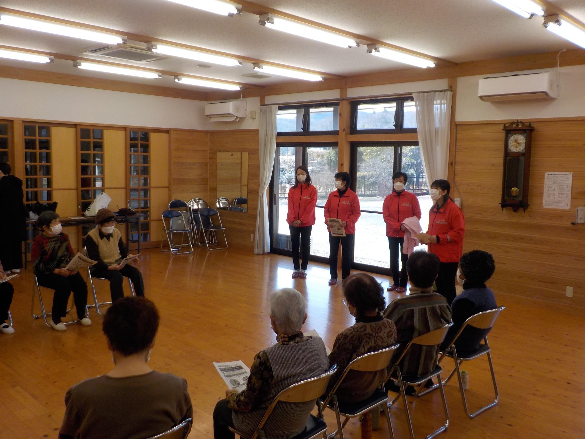 4人の赤いジャンパー姿の女性たちがヘルメットやたすきを手に立っていて、パイプ椅子に座った高齢女性の参加者の方々に説明している様子の写真