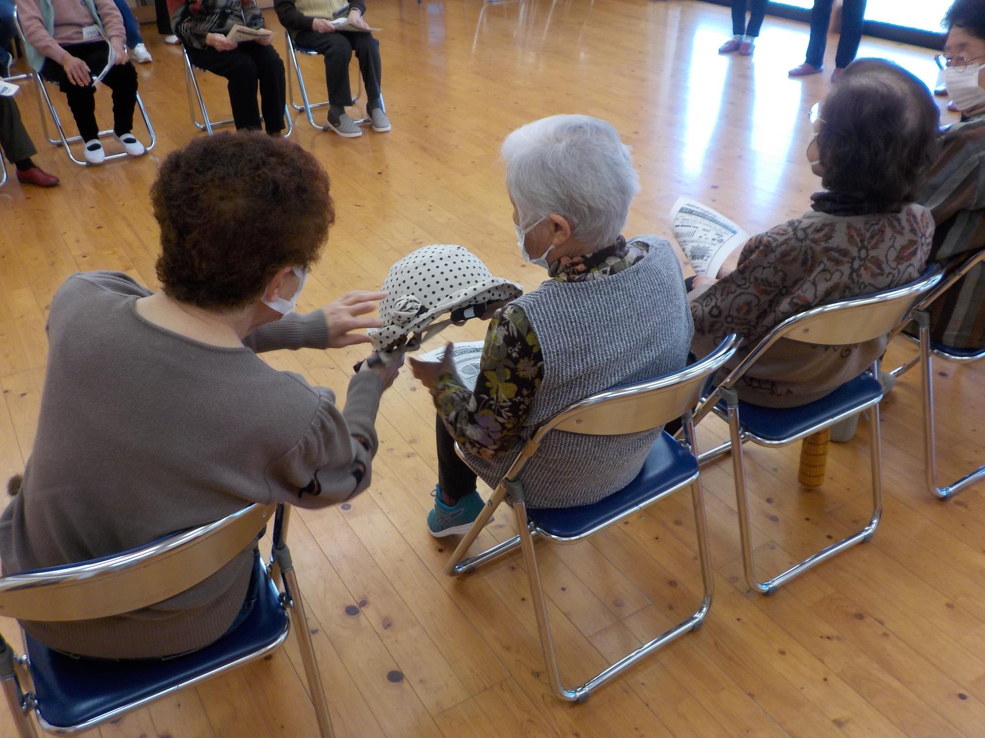 パイプ椅子に座って並ぶ3名の高齢女性の1人が隣の女性に水玉の帽子のようなヘルメットを手渡している様子の写真