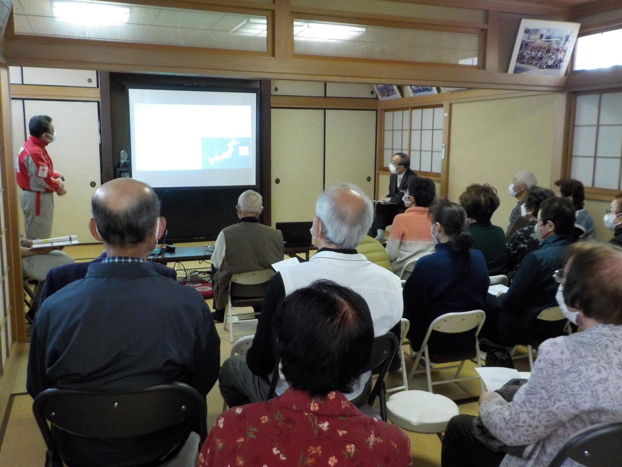 前方スクリーンの横で映像を見ながら話す吉田医師の話をパイプ椅子に座った多くの参加者の方々が聞いている様子を後方から撮影した写真