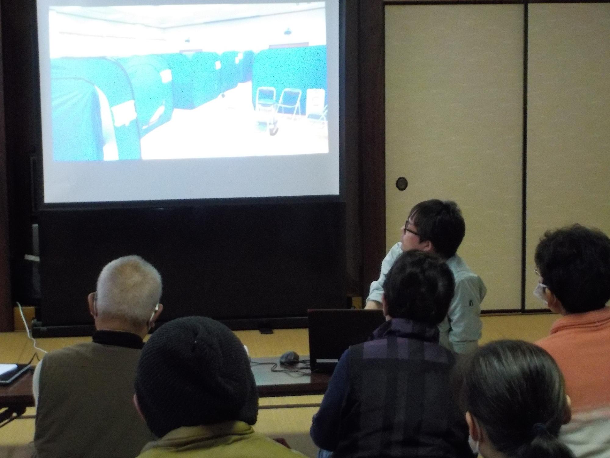 前方のスクリーンに映し出された避難生活の映像について説明する職員の話を視聴する参加者の方々の後姿を撮影した写真