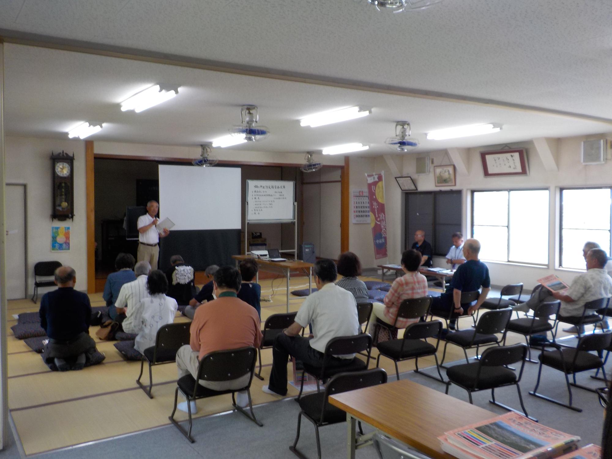 前方にホワイトボードとスクリーンがある畳敷きの会場で座布団や椅子に座り区長のあいさつを聞いている参加者の方々をななめ後方から撮影した写真