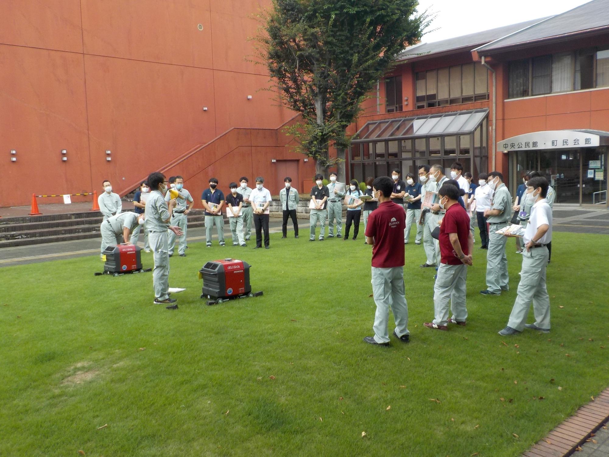 芝生の上に置かれた2台の赤い発電機の扱い方を男性が説明している様子を参加者たちが円になり聞いている写真