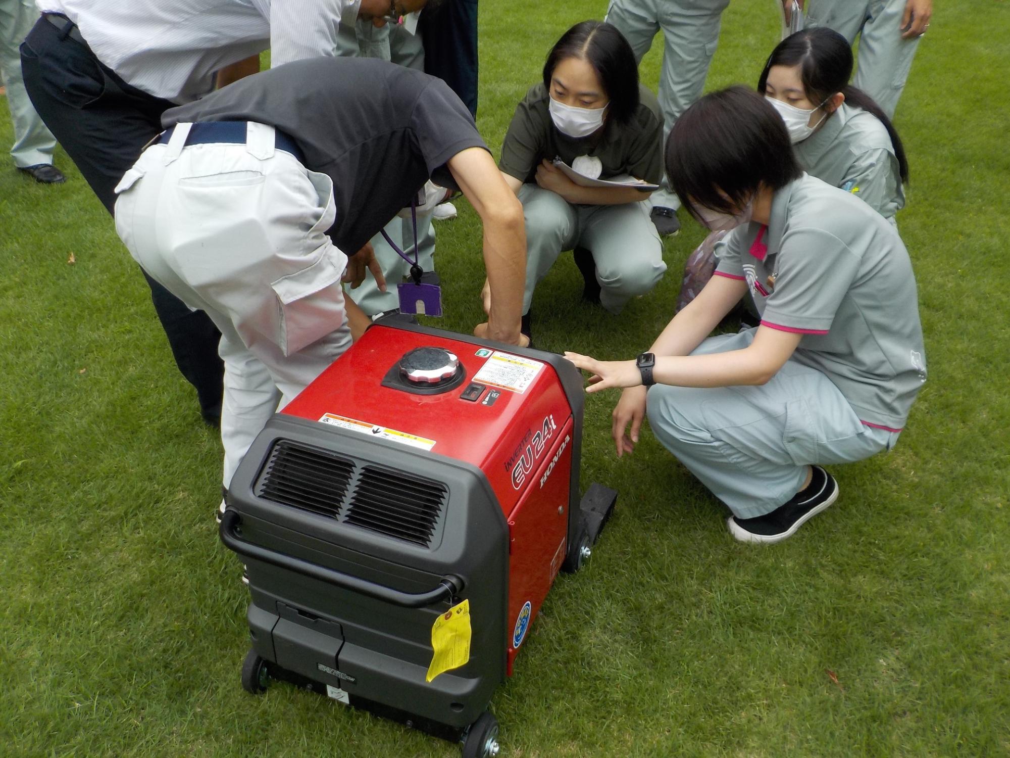 訓練の参加者たちが芝生の上に置かれた赤い発電機の扱い方を男性から教わっている写真