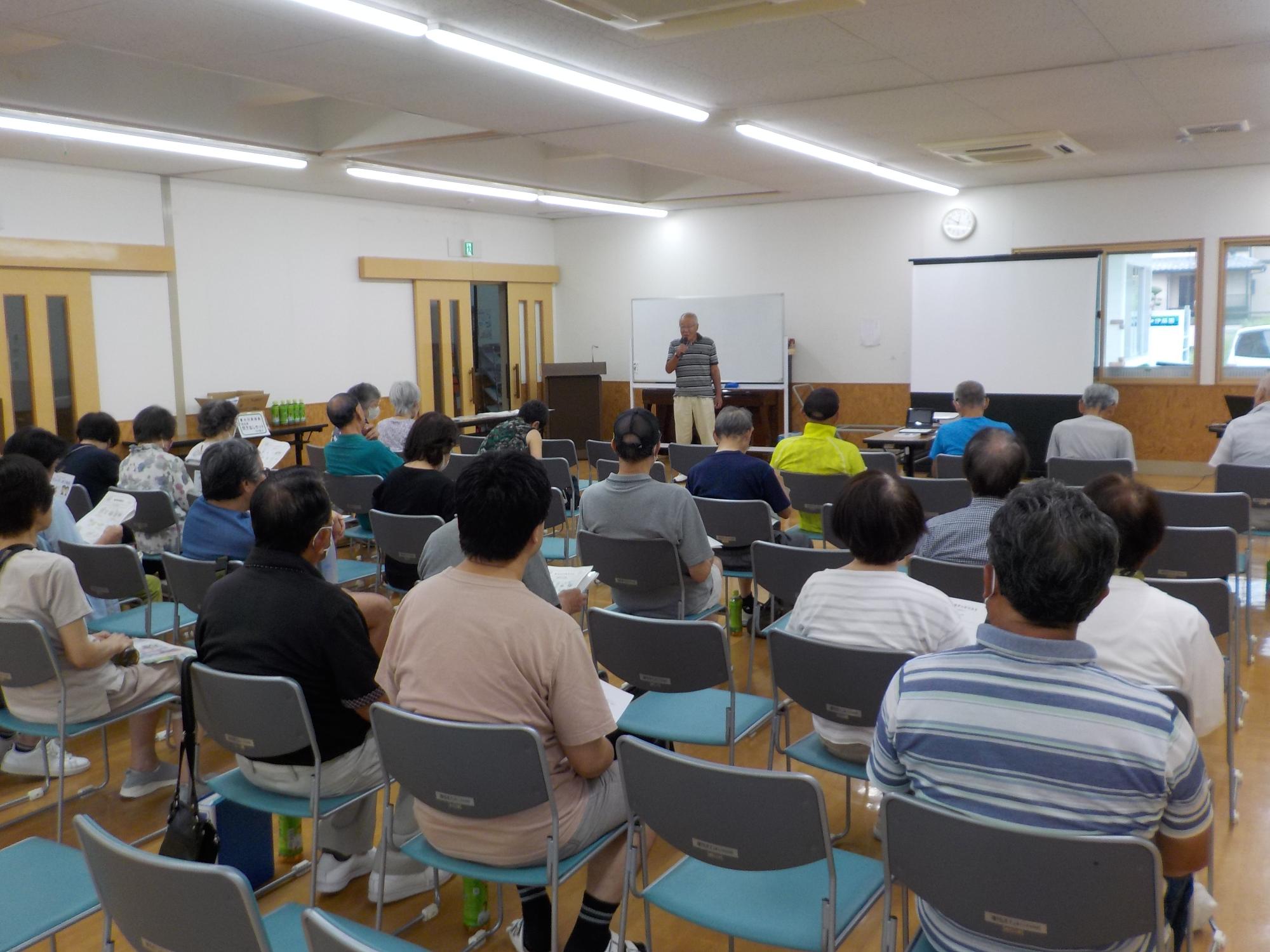 椅子に座っている参加者達の前に立ち、あいさつをしている石塚区長の写真