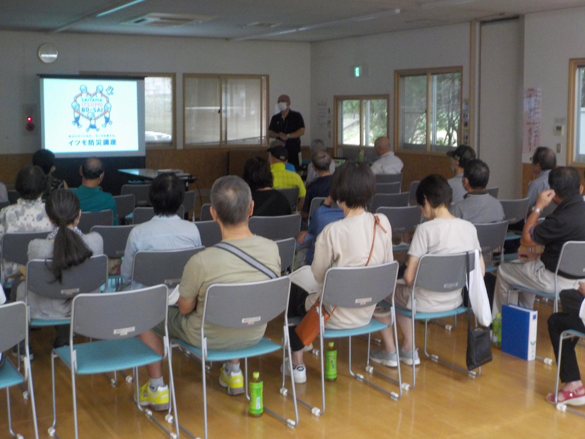 照明が落とされ薄暗くなった室内前方のスクリーンに映し出された資料を、椅子に座った参加者達が見ている様子を後方から撮影した写真