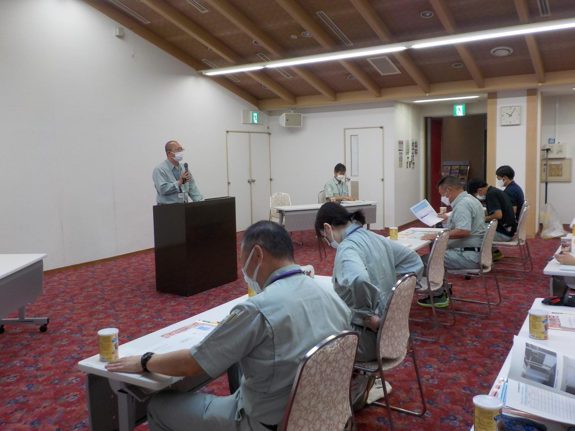 訓練の参加者たちが、会議室でマイクを持った男性の話を聞いている写真