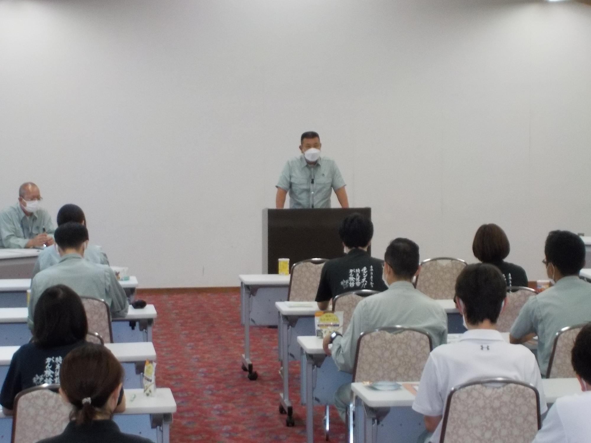 会議室の前の方にある講師用の机のところに立って話をする男性と、男性の話を聞く参加者たちの写真