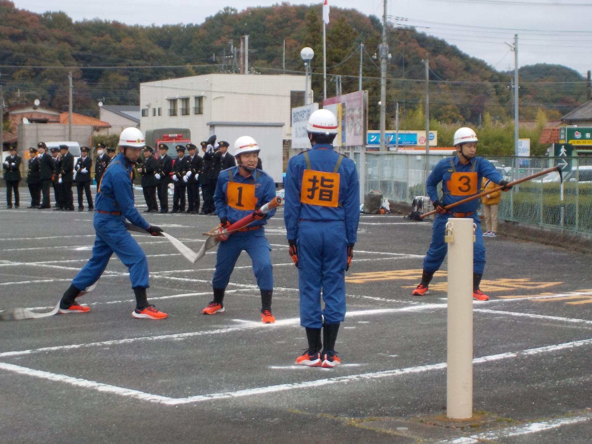 消防団員たちが広い駐車場で消防ポンプ操法の訓練を行っている写真