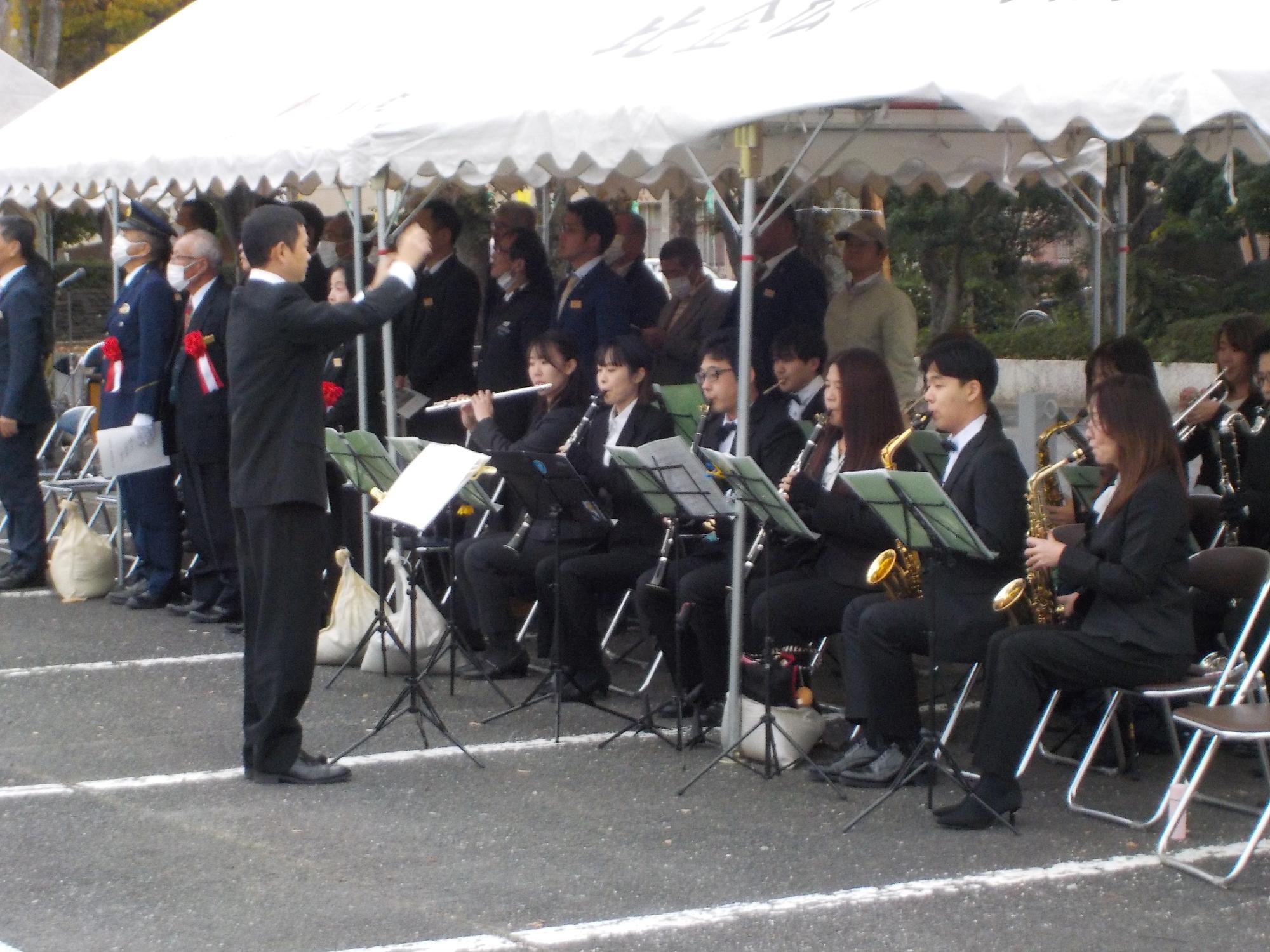 複数の男女が指揮者を前にして椅子に座りながら楽器を演奏している写真