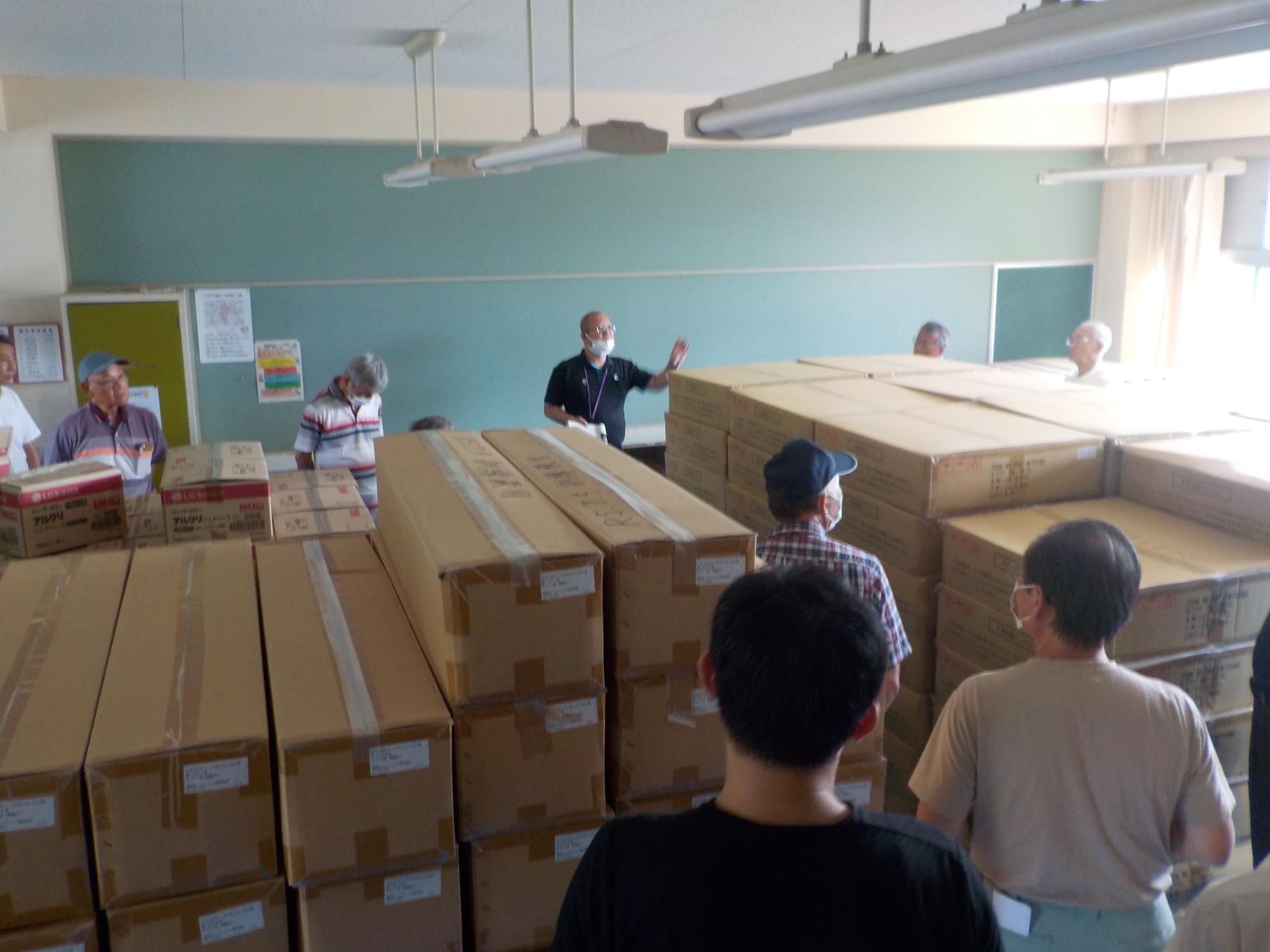 室内いっぱいに山積みに置かれた備蓄品の周りに参加者が集まり、室内前方で話す男性の説明を聞いている様子の写真