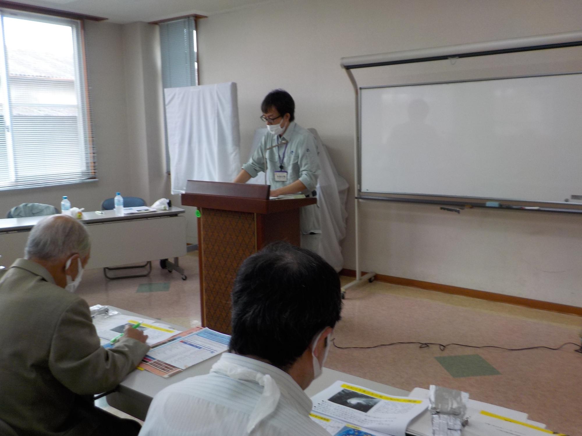 部屋の前の方にある講師用の机のところに立って話をする男性講師と、講師の話を聞く参加者たちの写真