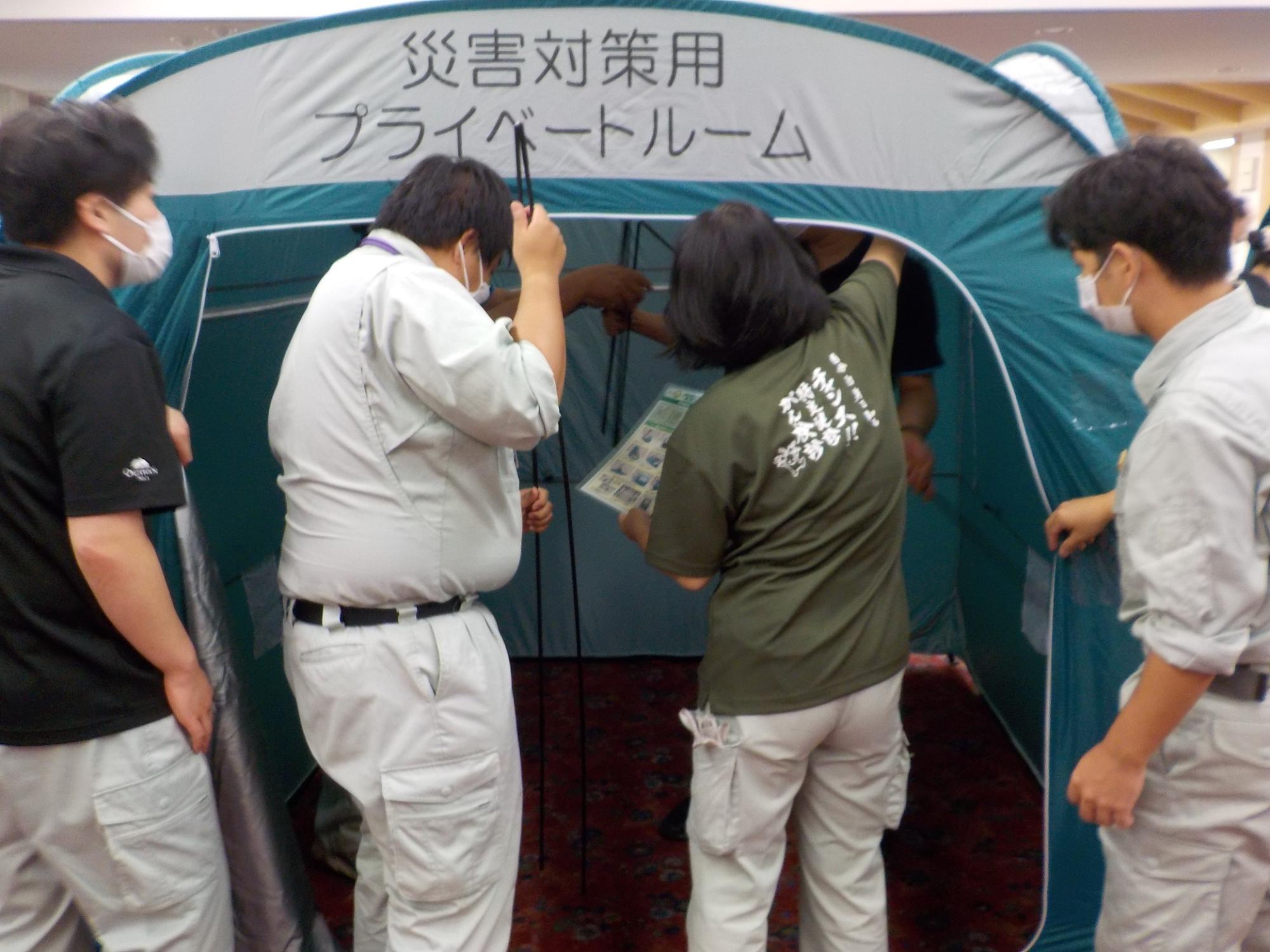 外観が組み立てられた災害対策用プライベートルームテントの内部を複数の方々で組み立てている写真