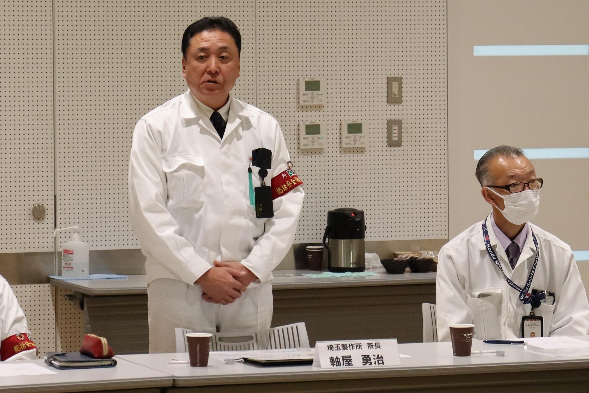 上下白い作業着を着た軸屋所長が両手を前で重ね挨拶をしている写真