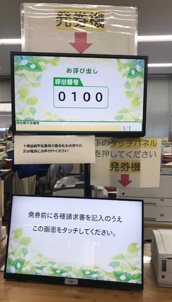 下矢印と「発券機」と書かれた表示の下のモニターに「お呼び出し【呼出番号】0100」と表示され、「下のタッチパネルを押してください」と書かれた表示の下のモニター「発券前に各種請求書を記入のうえこの画面をタッチしてください。」表示された待合表示モニターの写真