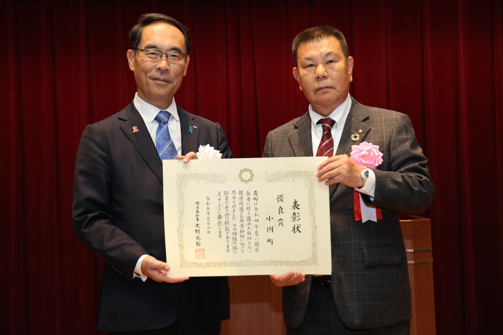スーツを着た二人の男性が、一枚の表彰状を一緒に両手で持って前に見せている写真