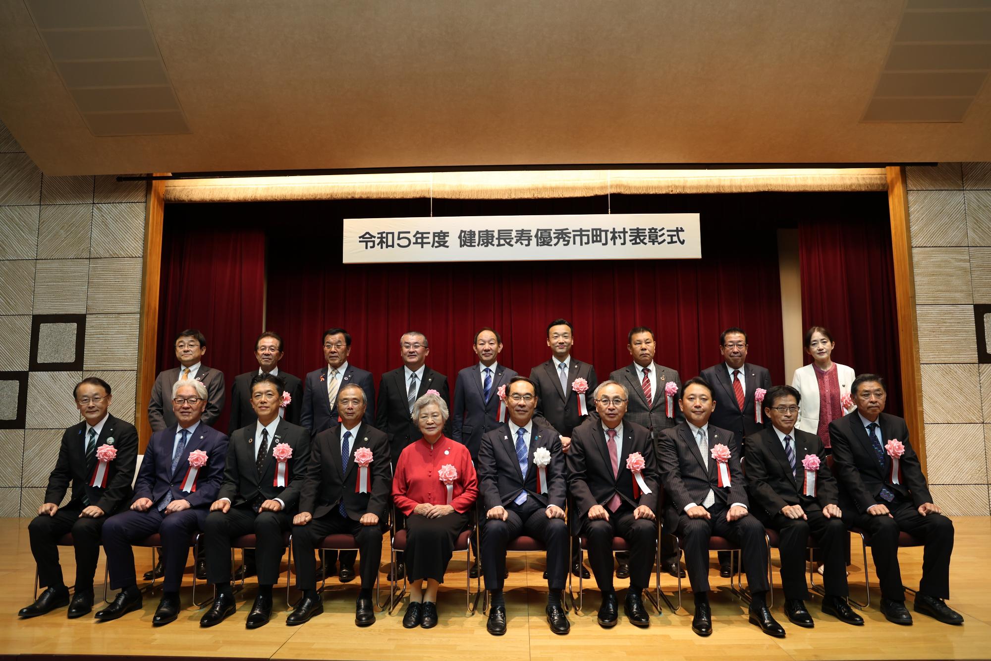 左胸に胸章リボンをつけた前列の椅子に10名座り、後列に立つ9名の受賞者が並んでいる記念写真