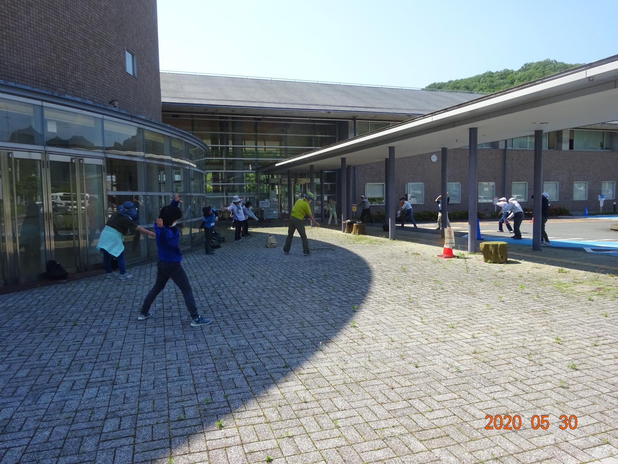 晴天の強い日差しを避けるように建物の日陰で出発前の準備体操をしている参加者の様子を後ろから撮影した写真