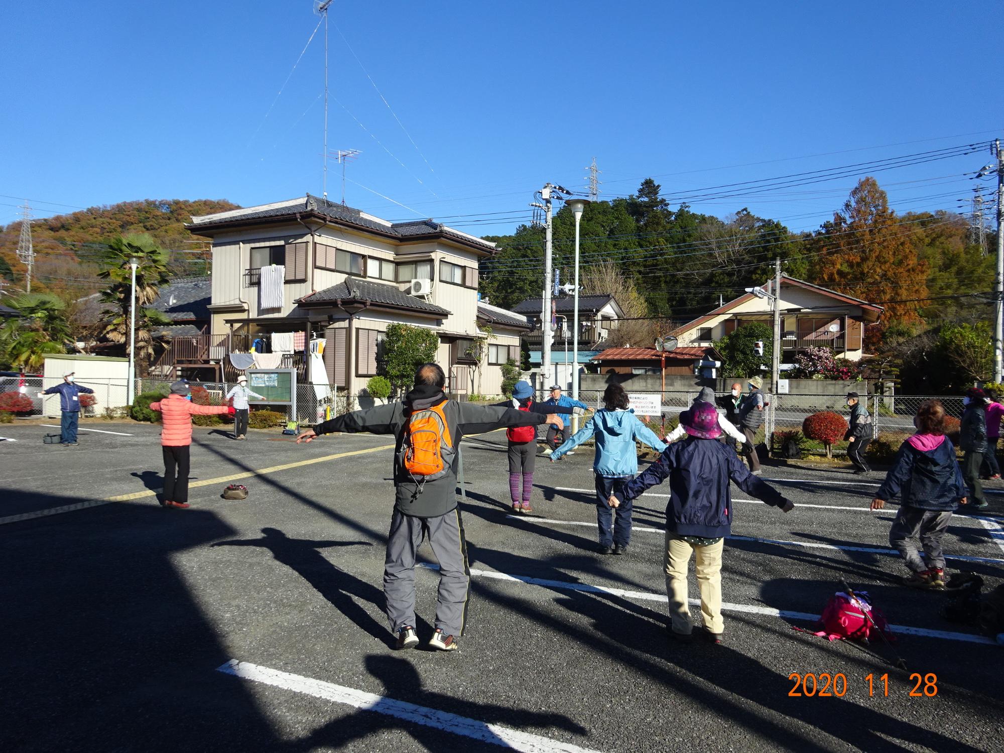 住宅地の前に広がる駐車場で参加者の方々が手を広げて体操をしている様子を後ろから撮影した写真