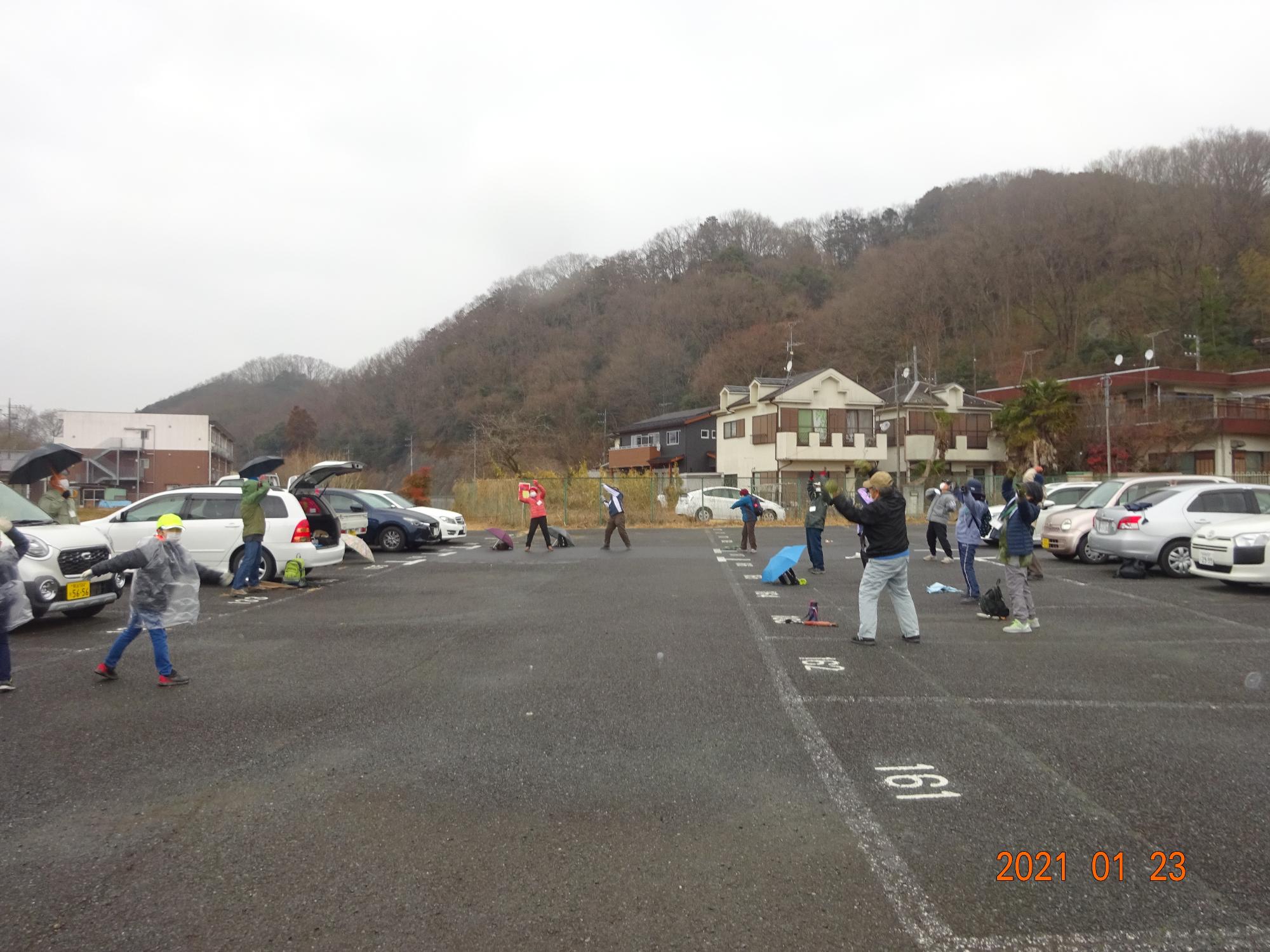 小雨の中19名の参加者の方々が駐車場で脇に傘を置いたりカッパを着たりして準備運動をしている様子を撮影した写真