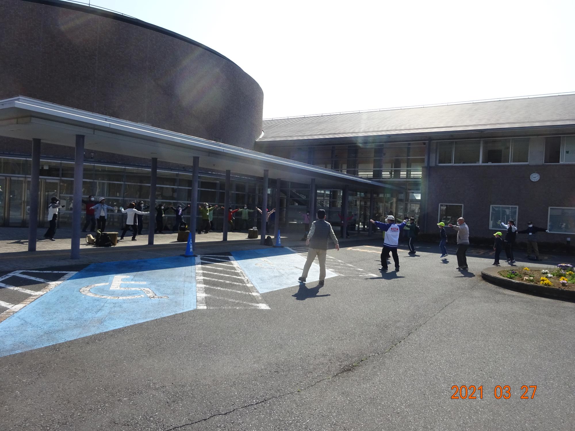 眩しい朝の光がさす中、建物の前と駐車場でたくさんの参加者の方々が準備運動をしている様子の写真