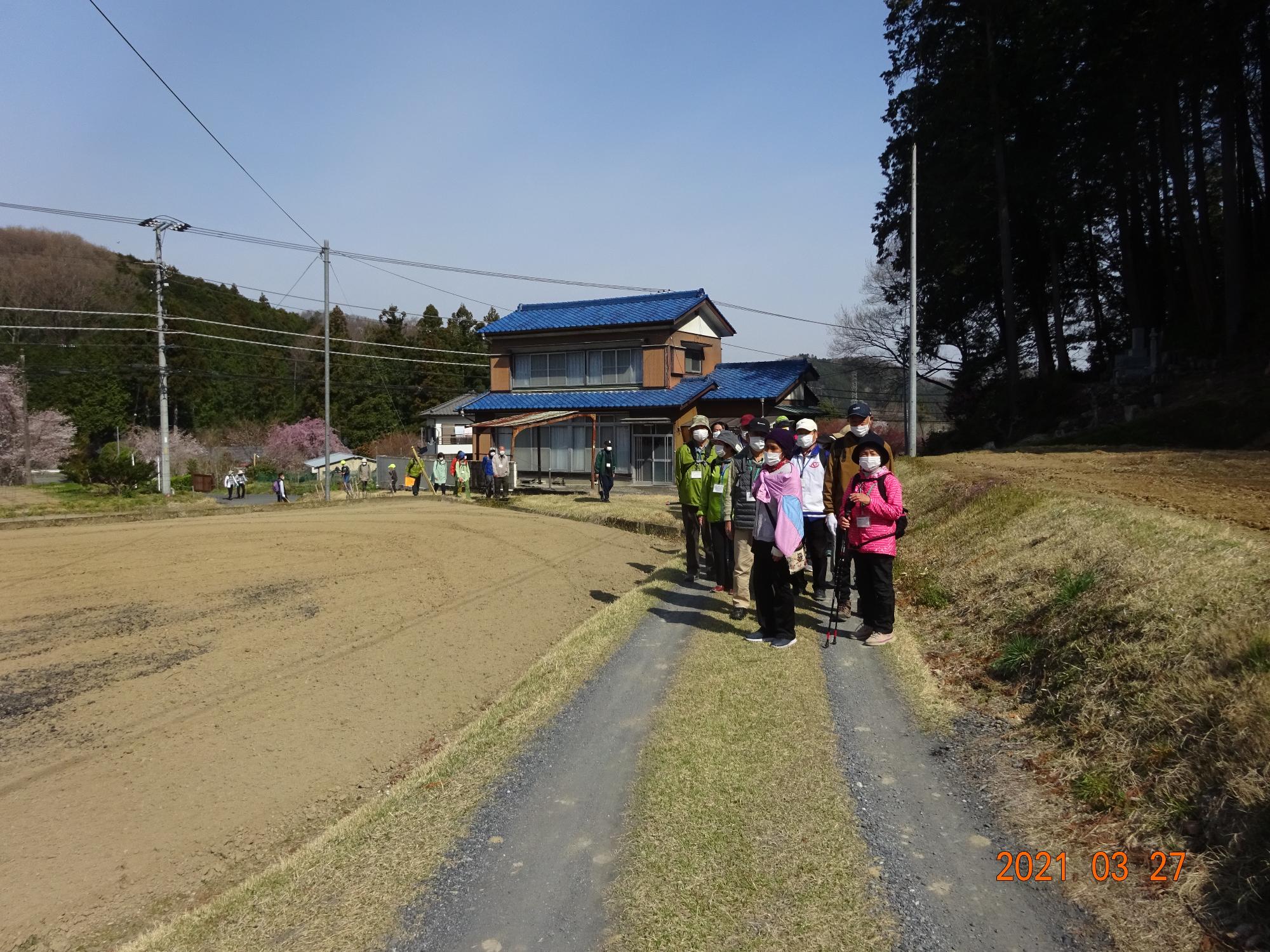後方の山には桜が咲いていて青い屋根の2階建てを背景にコースの途中で立ち止まっている参加者の方々を前方から撮影した写真