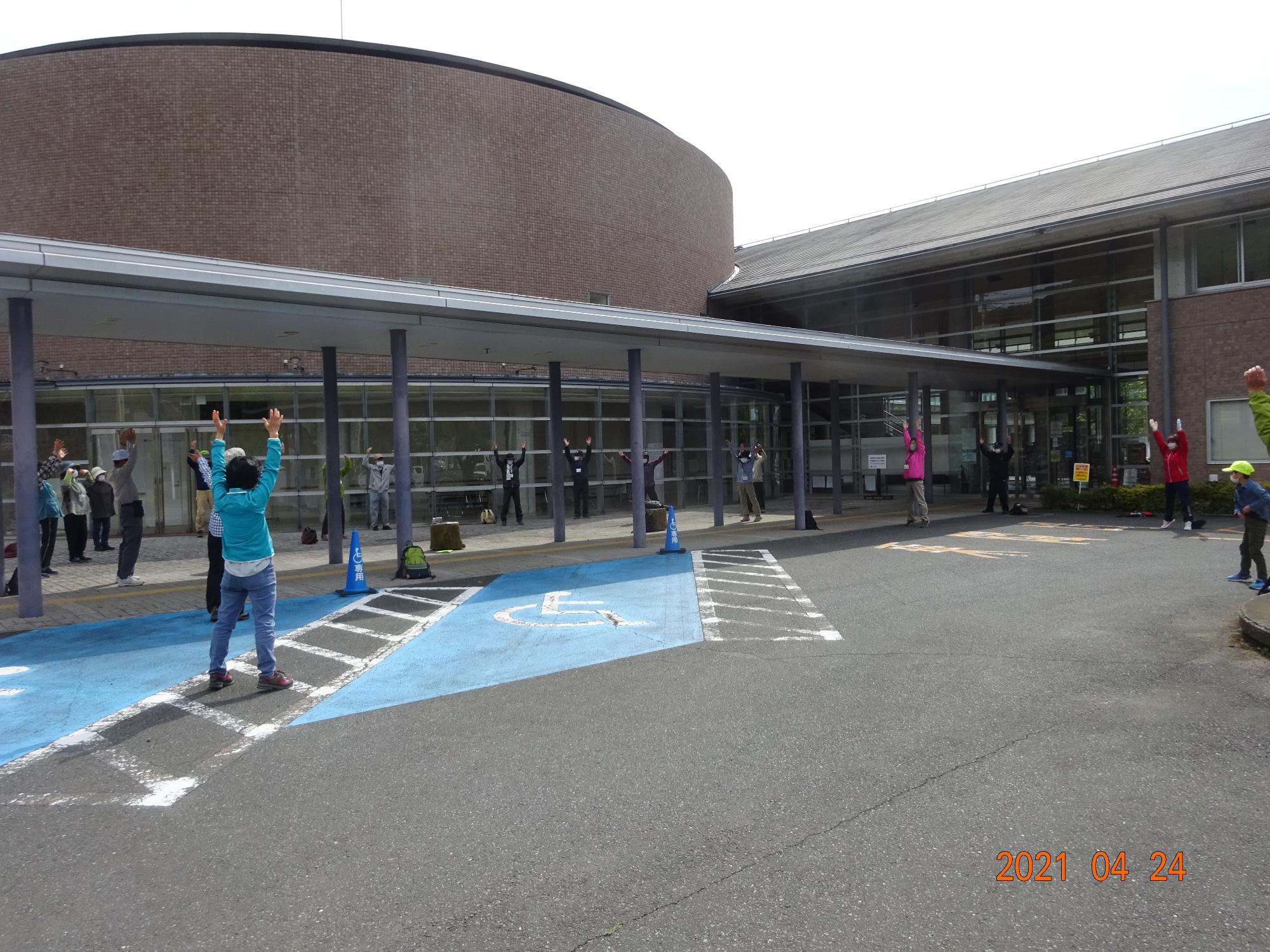 参加者たちが建物の前にあるスペースに広がりラジオ体操をしている写真