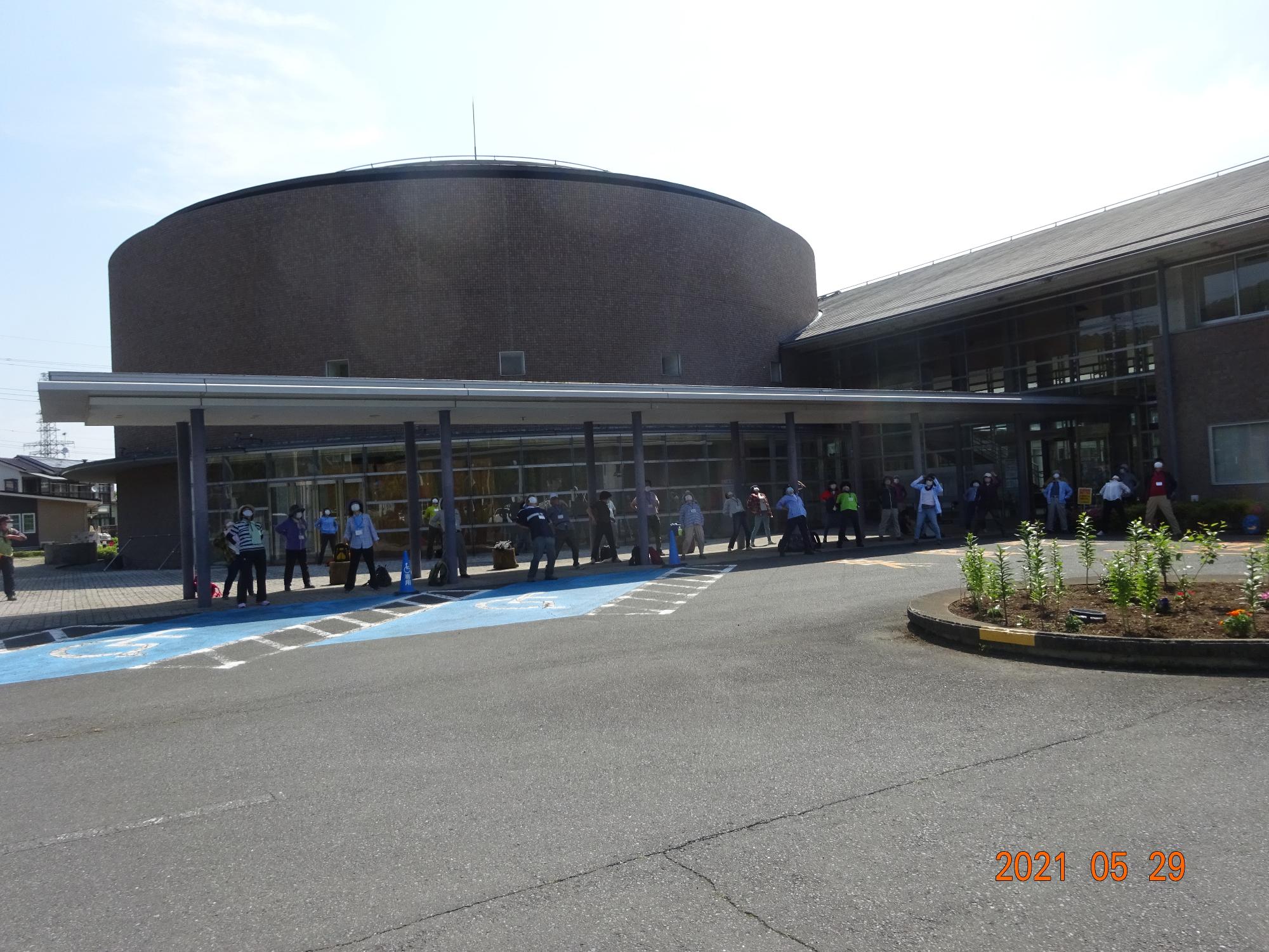 参加者たちが建物の前に設置された通路の雨除けの下で横並びになってラジオ体操をしている写真