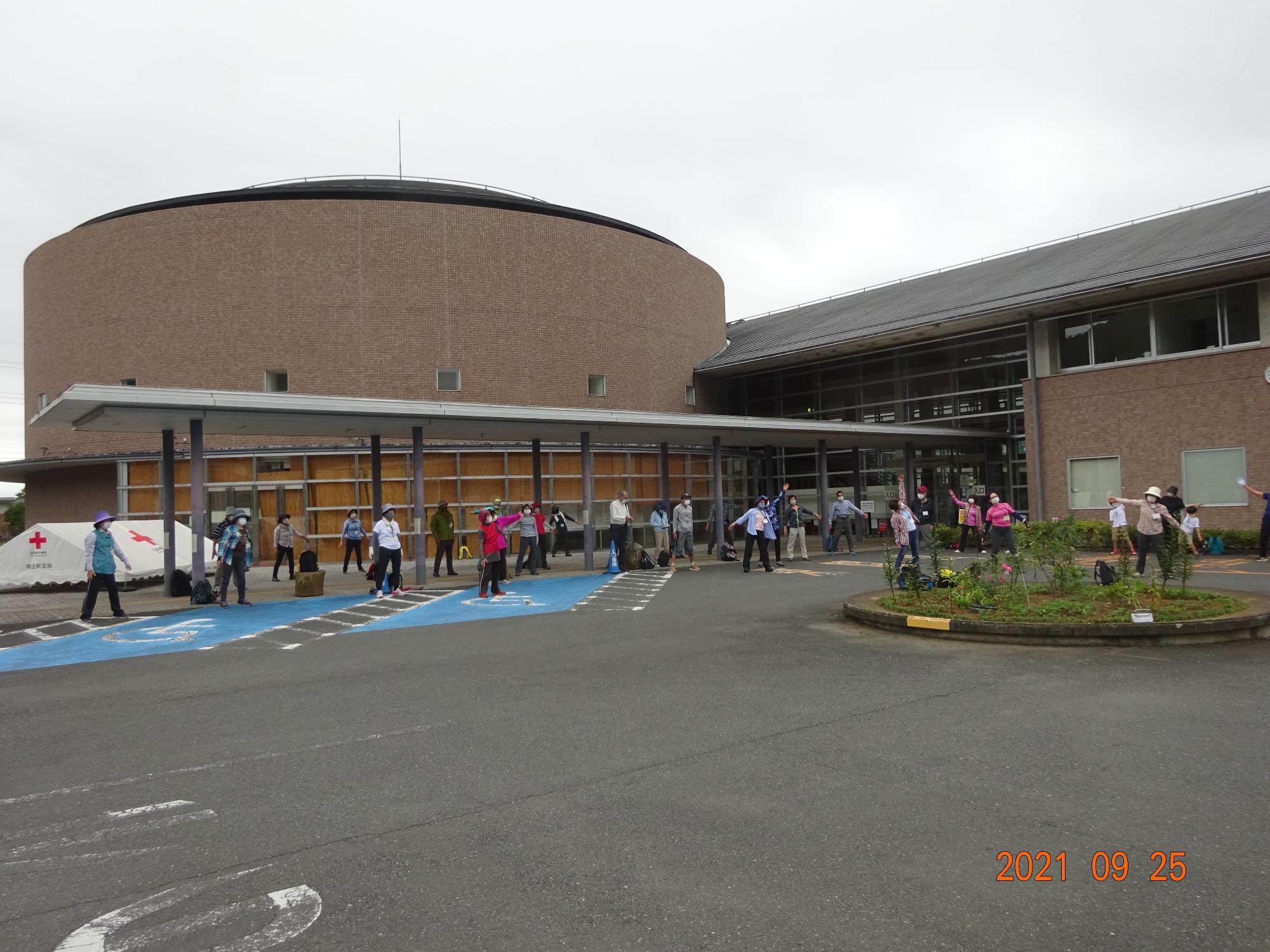 参加者たちが建物の前にある広いスペースの一角に広がってラジオ体操をしている写真