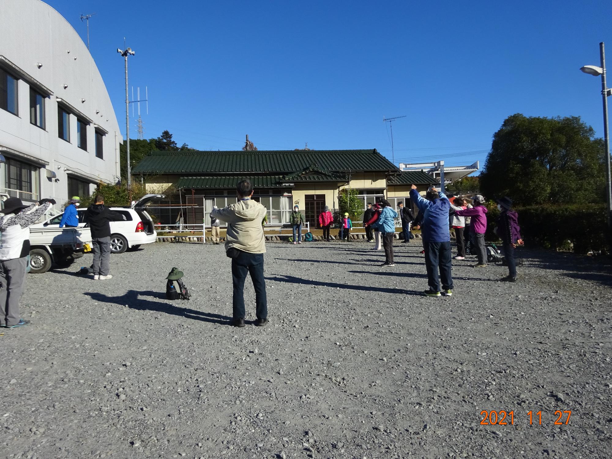 済んだ青空のもと、参加者たちが建物の前の広いスペースでラジオ体操をしている写真