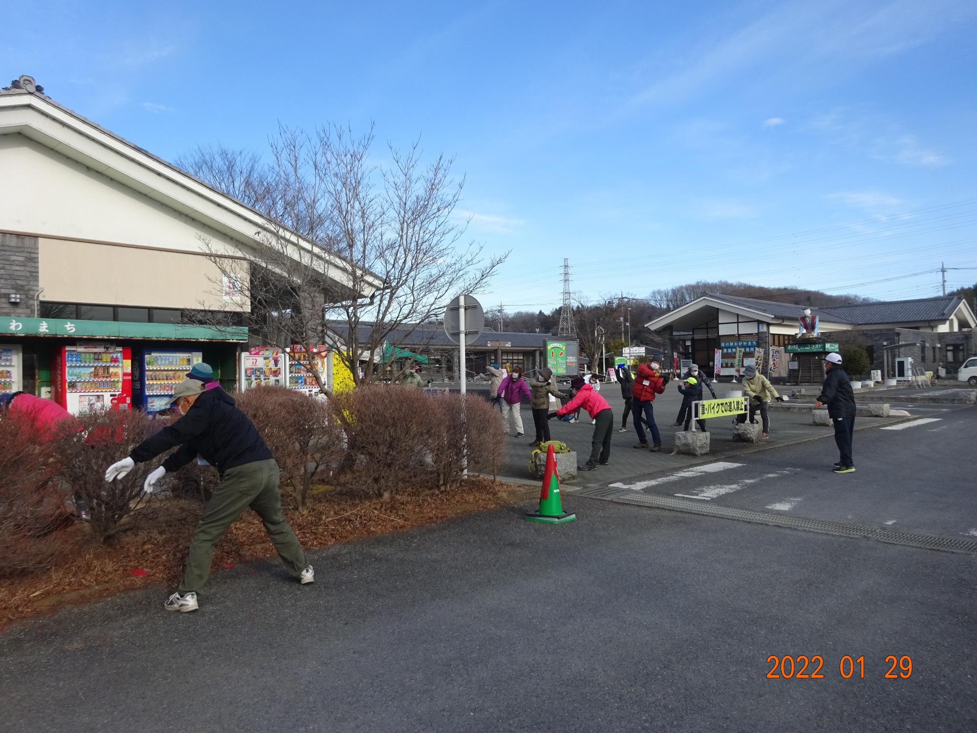 青空のもと、参加者たちが建物の前の広いスペースに広がりラジオ体操をしている写真
